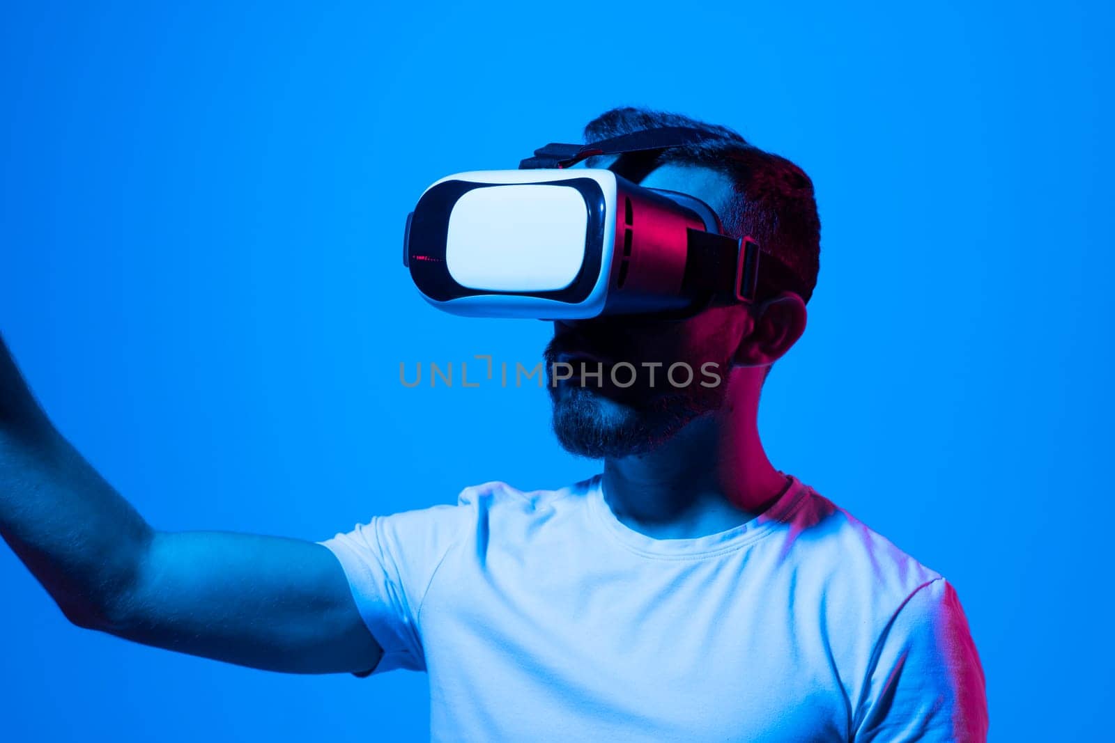 Handsome Bearded man in white t-shirt and VR headset enjoying experience playing a game and gesturing while standing against wall in neon light. by vovsht