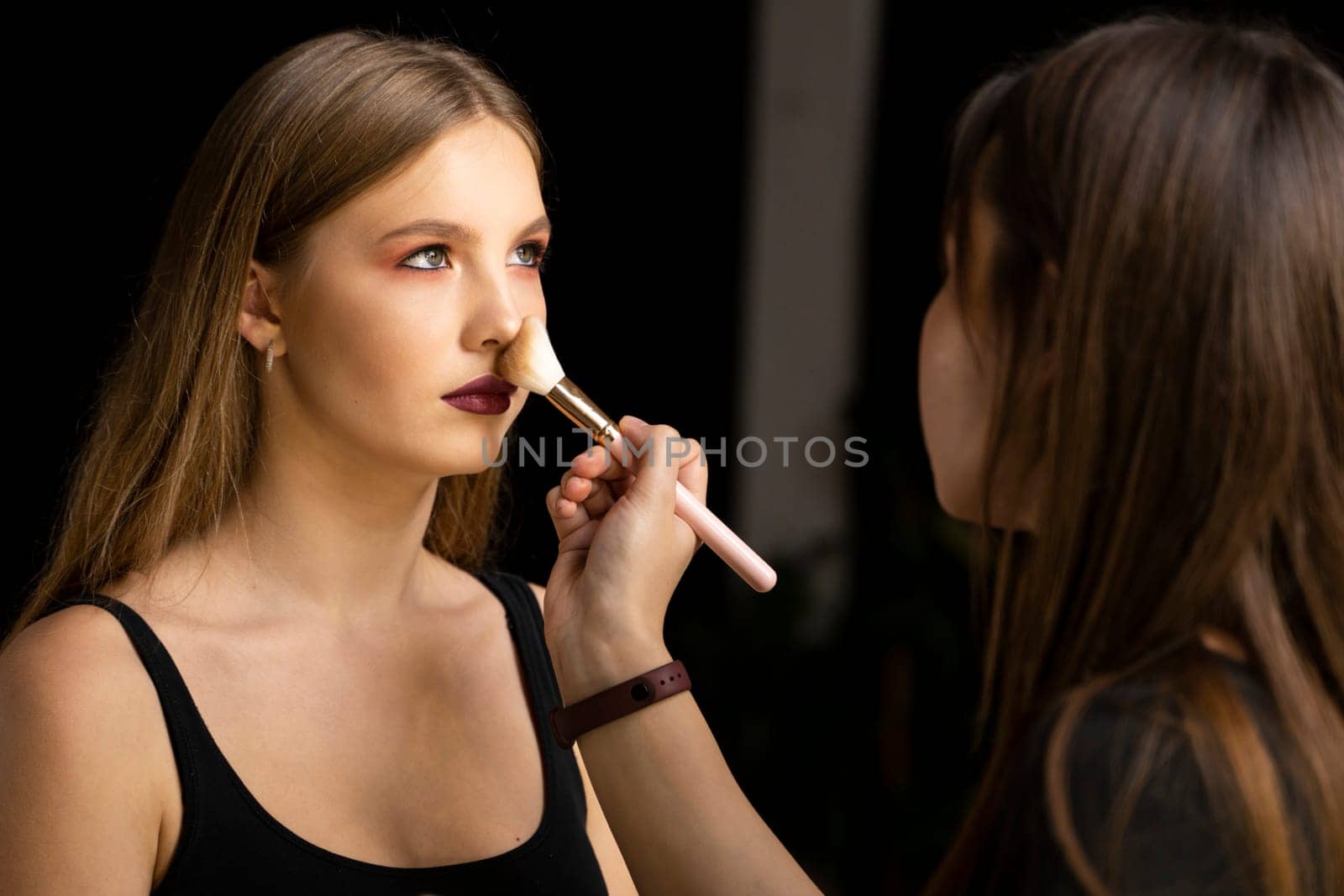 Professional make-up artist working with brush on model face. Process of making makeup. Portrait of young blonde woman in beauty salon interior. Applying tone to skin