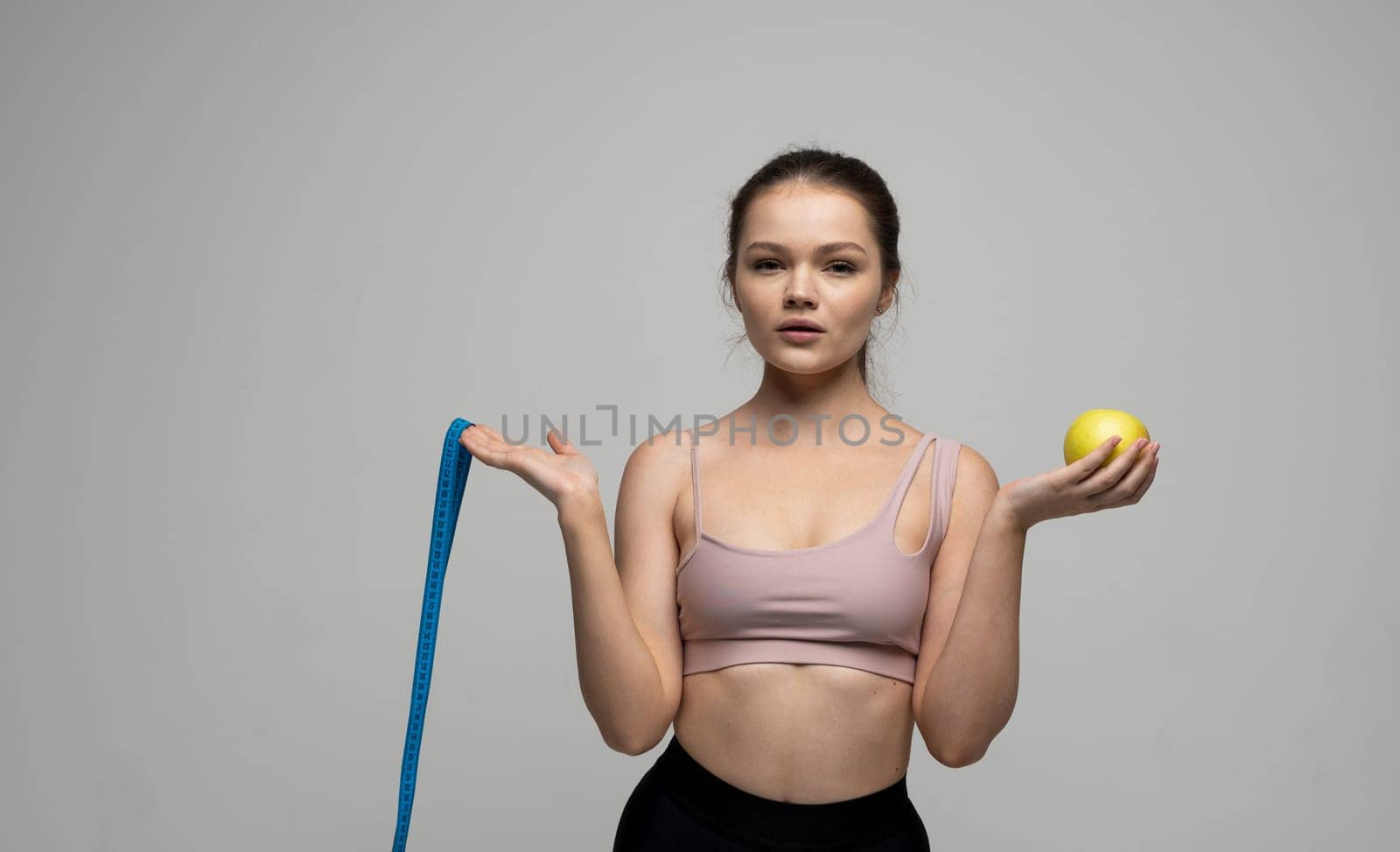 Confused attractive sexy brunette women in sport outfit standing with a measuring tape and holding a apple on a white background. Beauty and health concept. Healthy eating and diet. by vovsht