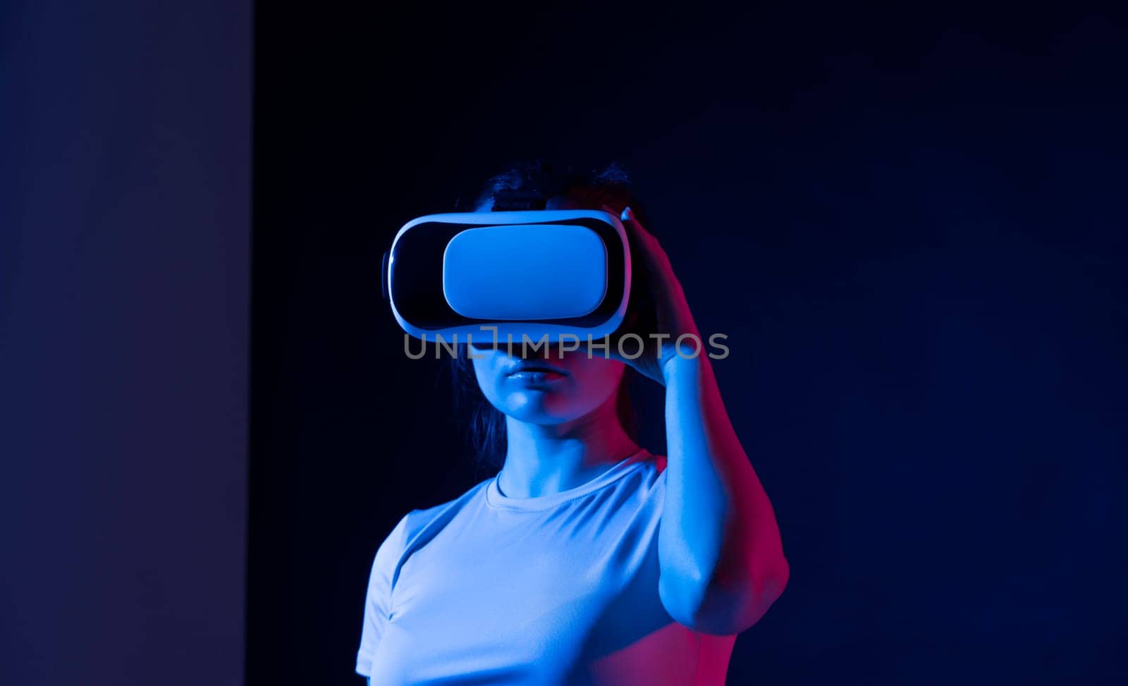 Young brunette woman with raised hands using virtual reality goggles at home in neon light. Modern future technology. by vovsht