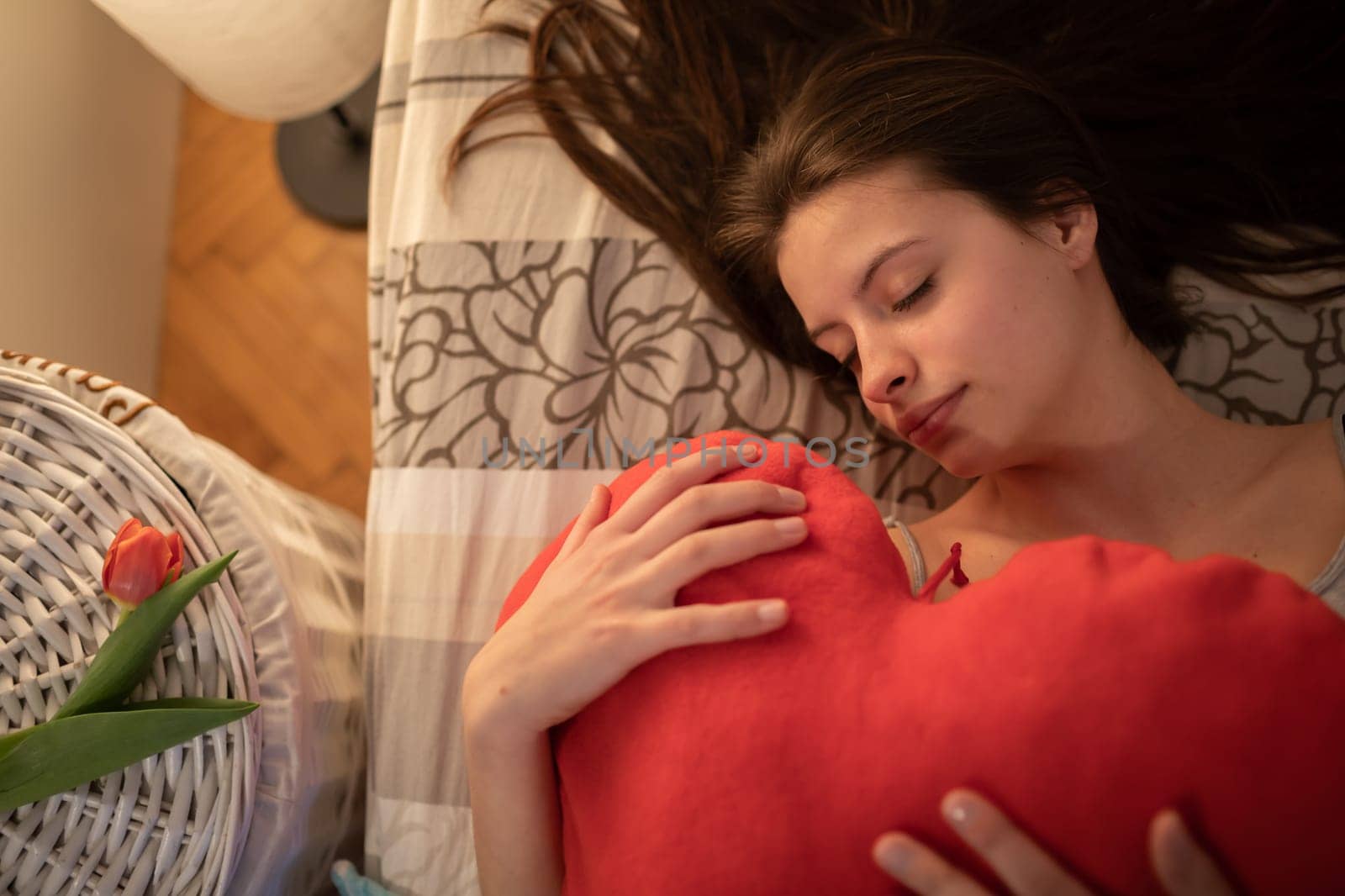 The teenager fell asleep contentedly hugging a red heart-shaped pillow. The lovers' day passed nicely. The girl sleeps with the bedside lamp lit.