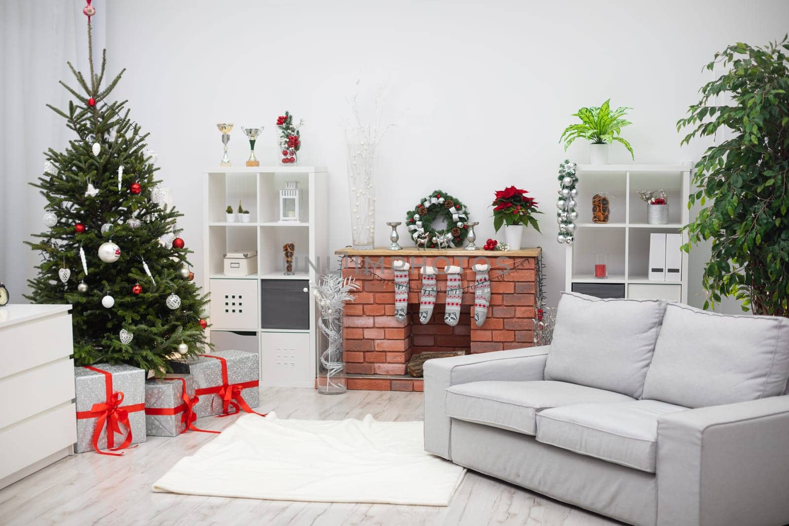 A bright living room with a fireplace. Live Christmas tree decorated in white and red. Silver gifts with red ribbons.