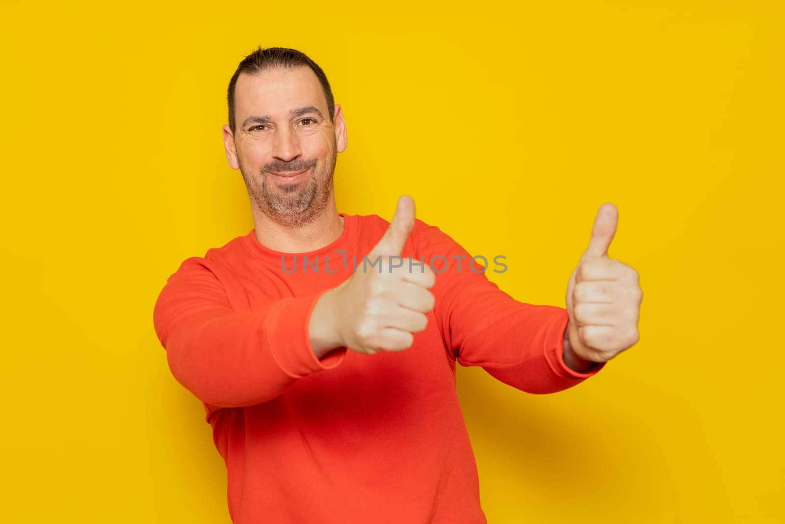 Portrait of a cheerful bearded hispanic man in red sweatshirt smiling and showing thumbs up to camera isolated over yellow background