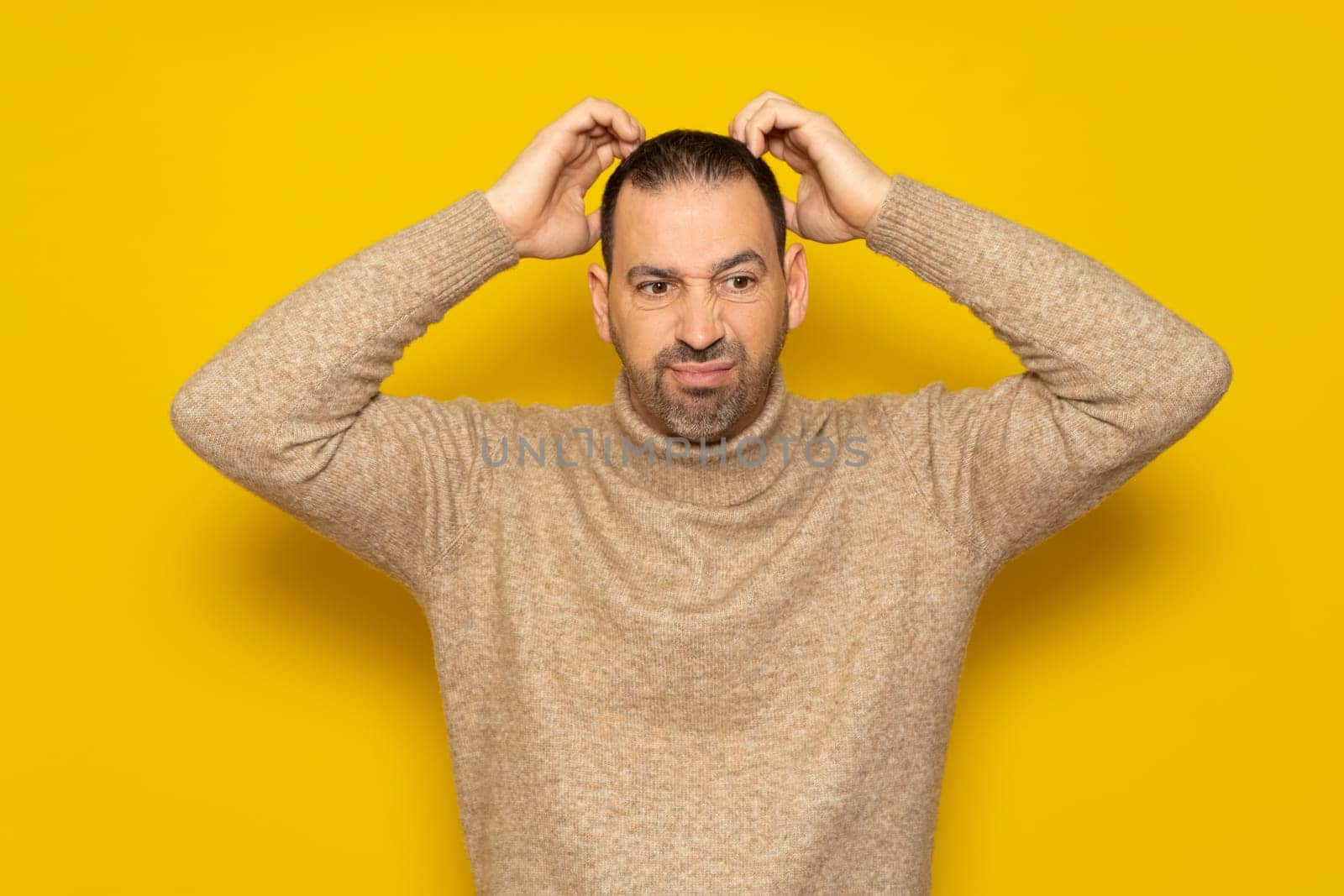 Bearded Hispanic man wearing a beige turtleneck scratching his head energetically while trying to solve a problem or doubt, isolated over yellow background