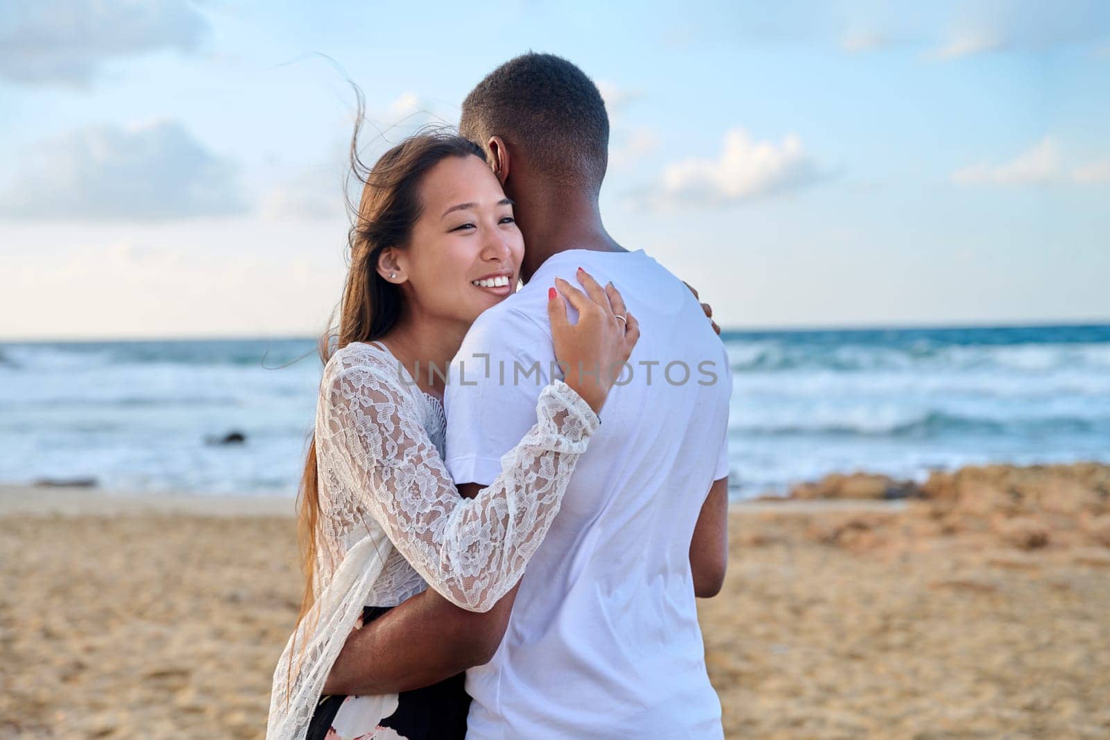 Young loving couple hugging on the beach. Face of beautiful Asian woman, man back. Love, relationship, honeymoon, vacation together, happiness, romantic, travel, family concept