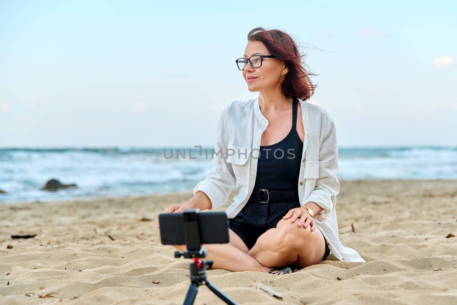 Middle aged woman sitting on beach with smartphone using video call. Female recording blog, vlog, talking online. Technology, telecommunications, lifestyle, freelance, training and business concept