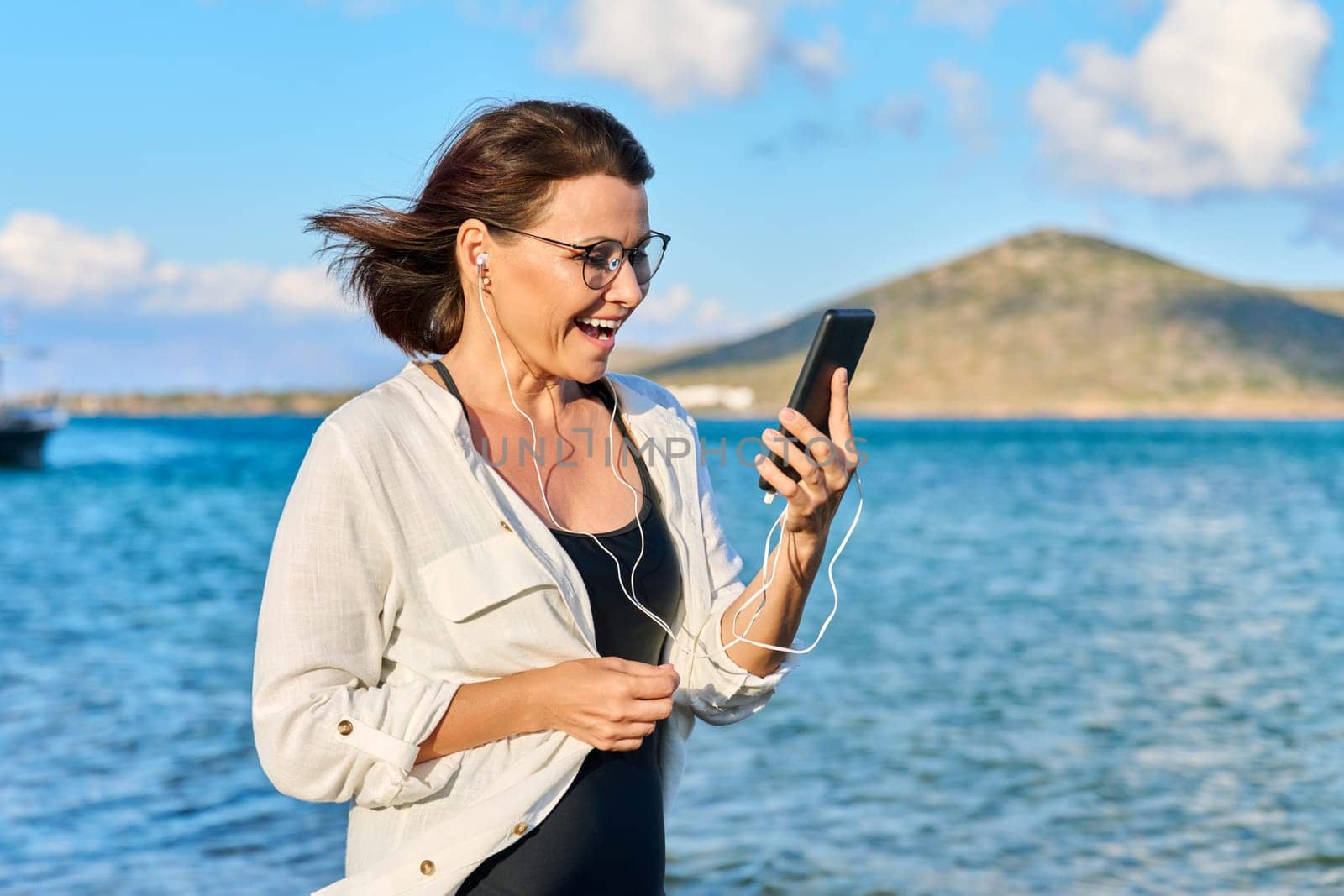 Happy 40s age woman in headphones with smartphone, sky sea nature. Beautiful female listening looking at smartphone screen walking relaxing on beach, copy space. Tourism, travel, vacation, technology