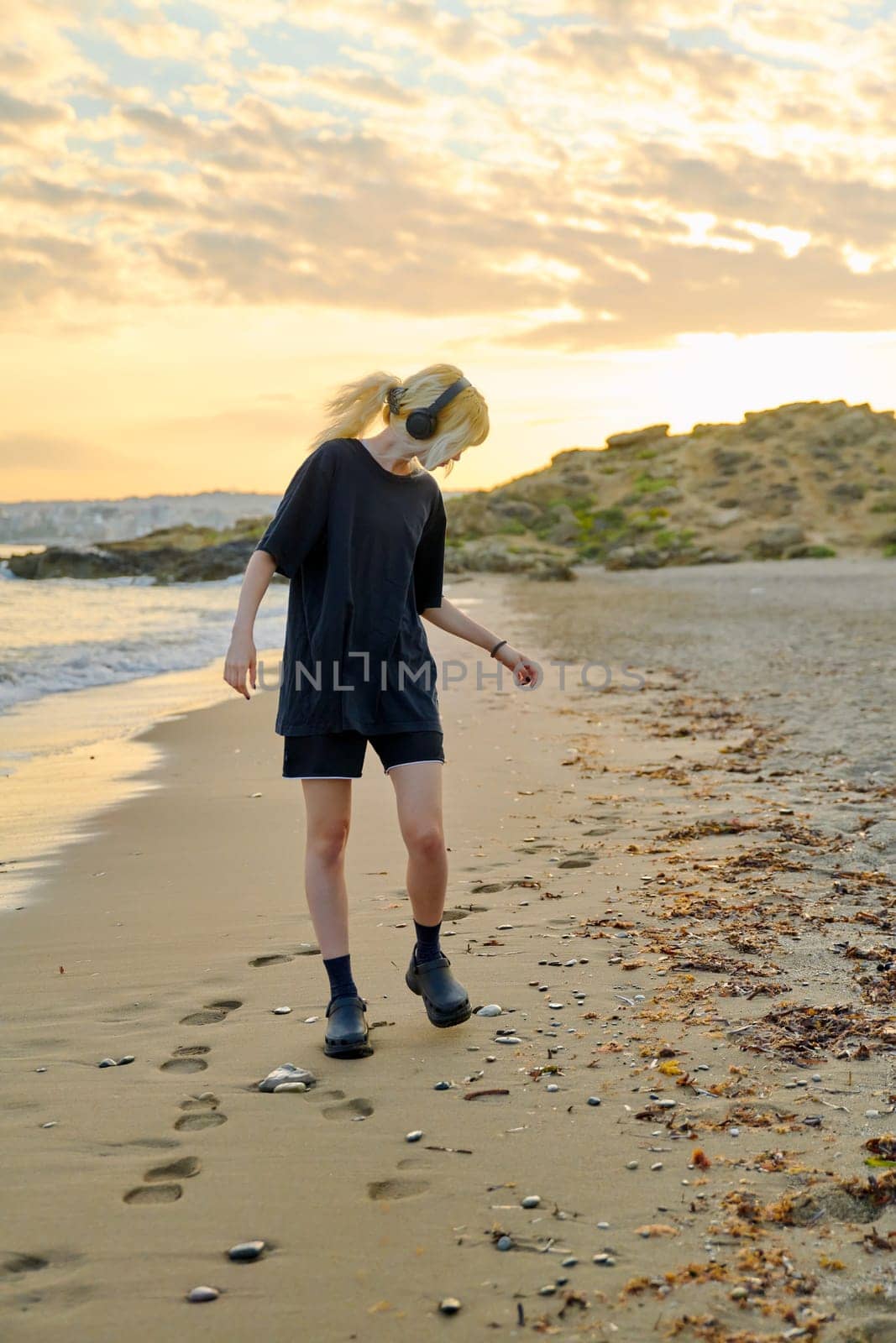 Young teenage female on beach in morning. Hipster teenager in headphones walking along seashore with picturesque colorful sea nature. Summer sunrise, freedom vacation beauty youth concept