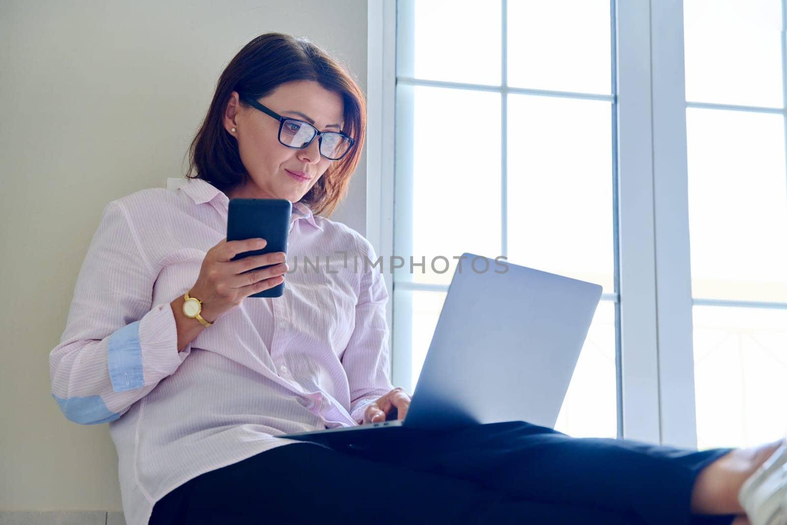 Business mature woman sitting on floor at home using laptop and smartphone. Remote business, work, technology, 40s people concept