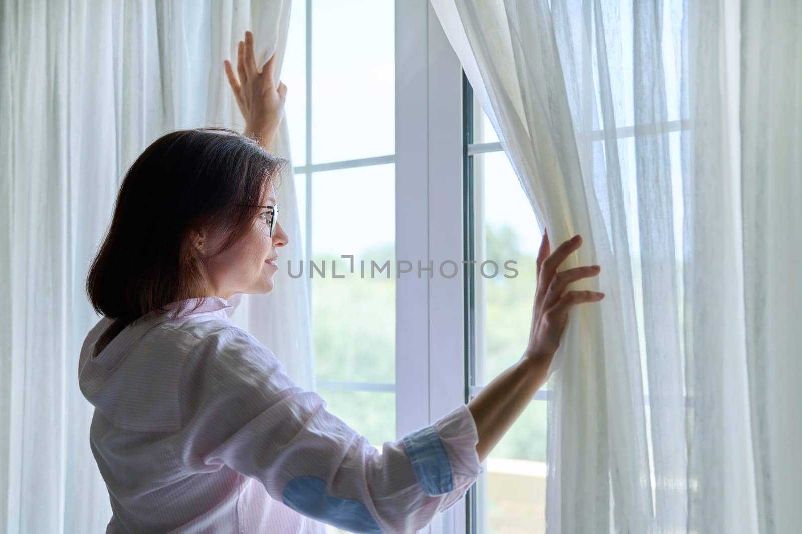 Smiling positive mature woman looking out the window. Happy 40s female standing near window at home