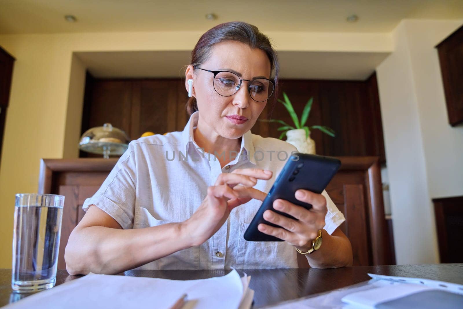 Face of mature woman looking at webcam, talking, at her desk at home. Middle-aged female in headphones with smartphone in hands talking online, remote business, virtual meeting, consultation, training