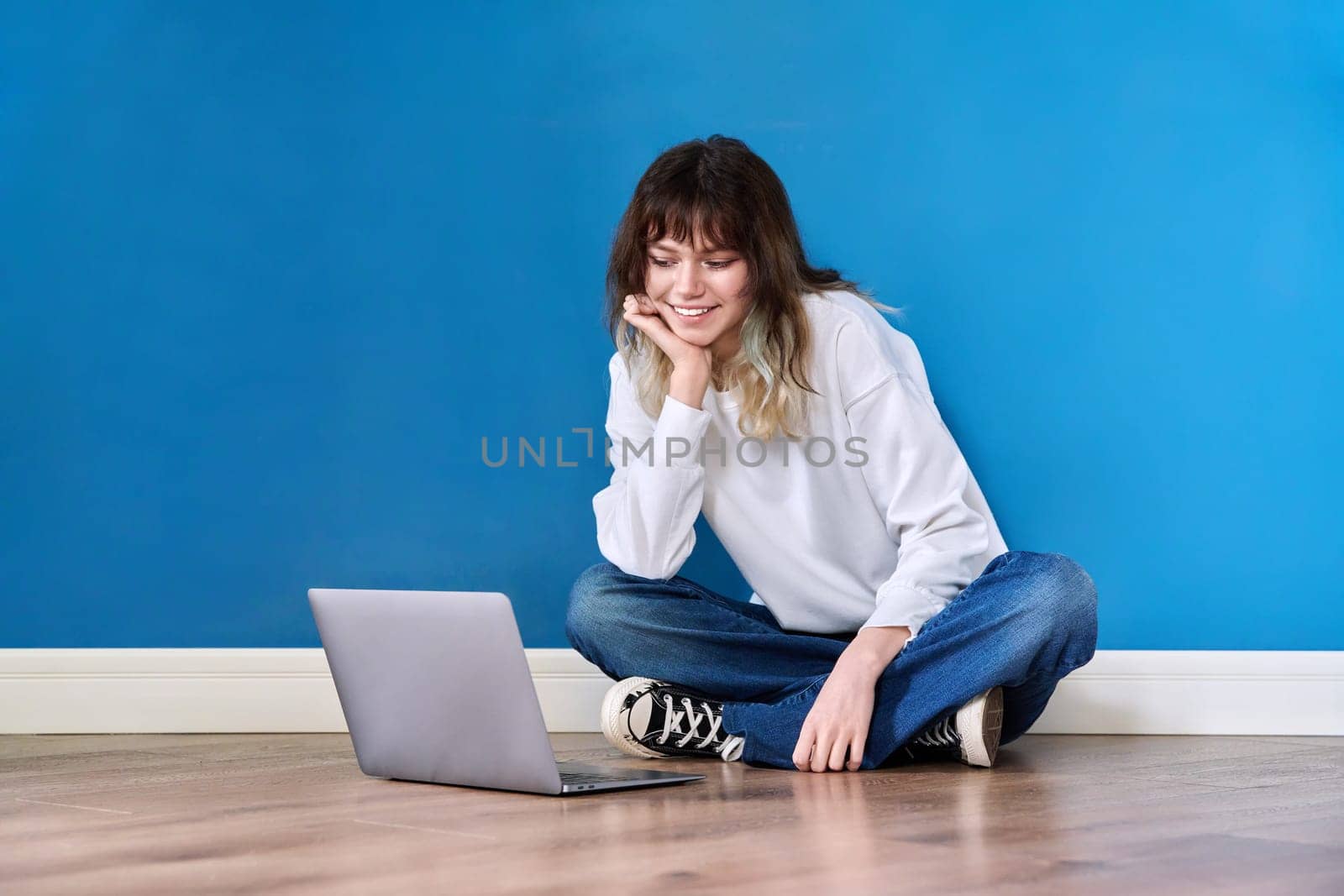 Beautiful teenage female sitting on floor with laptop on blue background. Hipster teenager looking at laptop screen, copy space wall. Youth, lifestyle, leisure, education, technology concept
