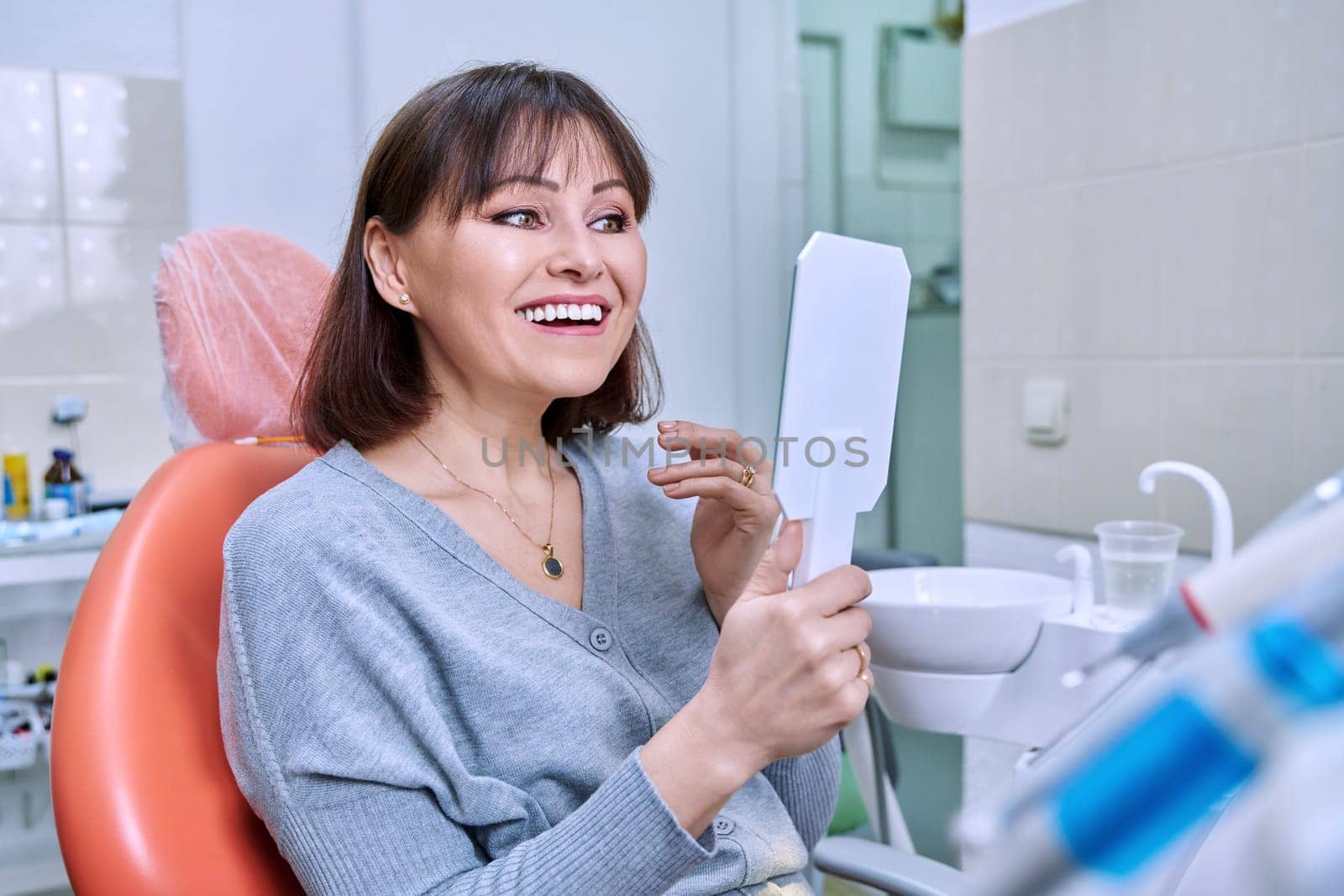 Smiling middle aged woman in dental chair in dentist's office with mirror looking at her teeth. Treatment, therapy, dental care, prosthetics, dentistry concept