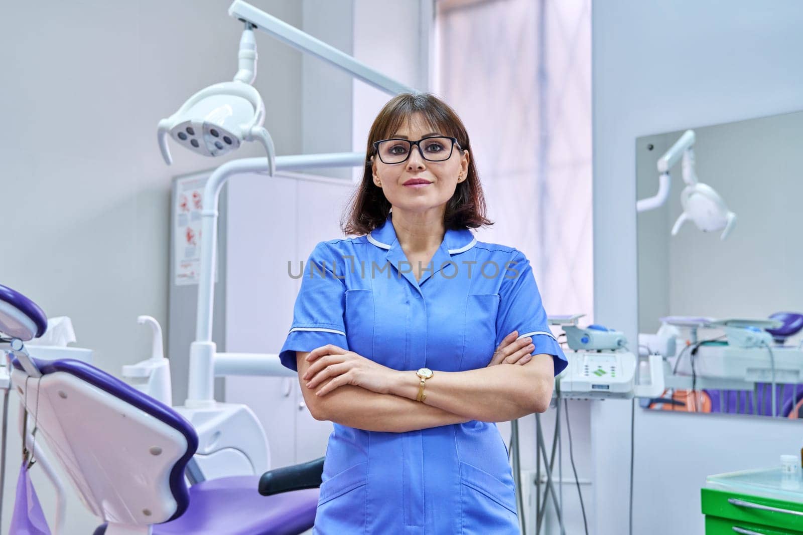 Portrait of smiling nurse looking at camera in dentistry. Confident female with folded hands, dental clinic equipment background. Dentistry, medicine, health care, profession, stomatology concept