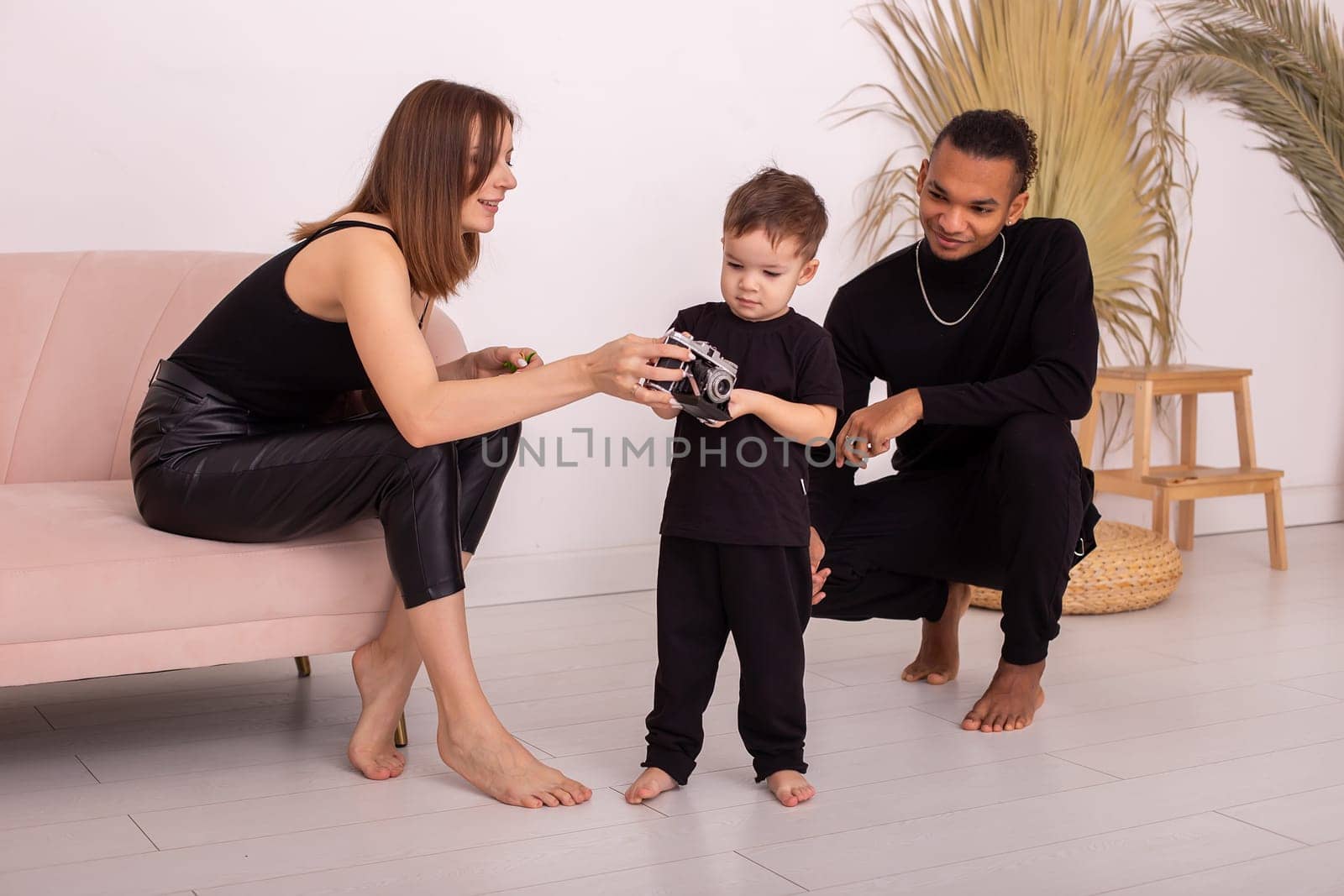 Happy multiracial family with little boy, in black clothes, in the light living room, playing , hold old camera
