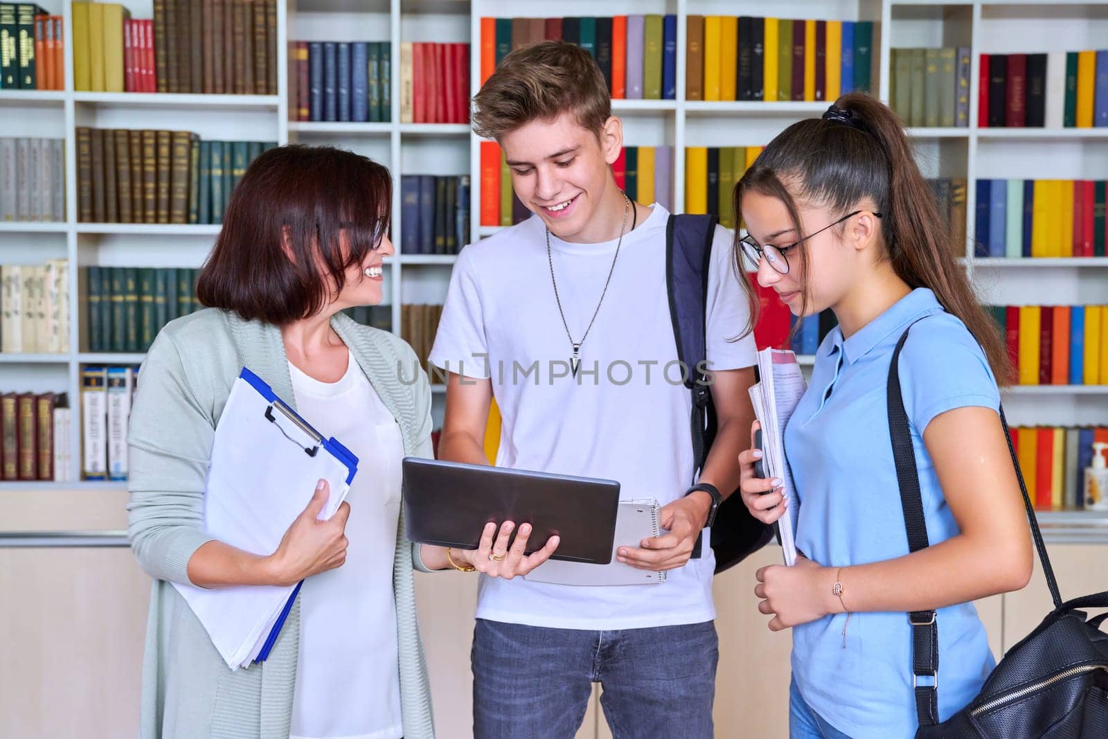 Students teenagers talking with woman teacher mentor, library shelving with books background. High school, education, knowledge, adolescence concept