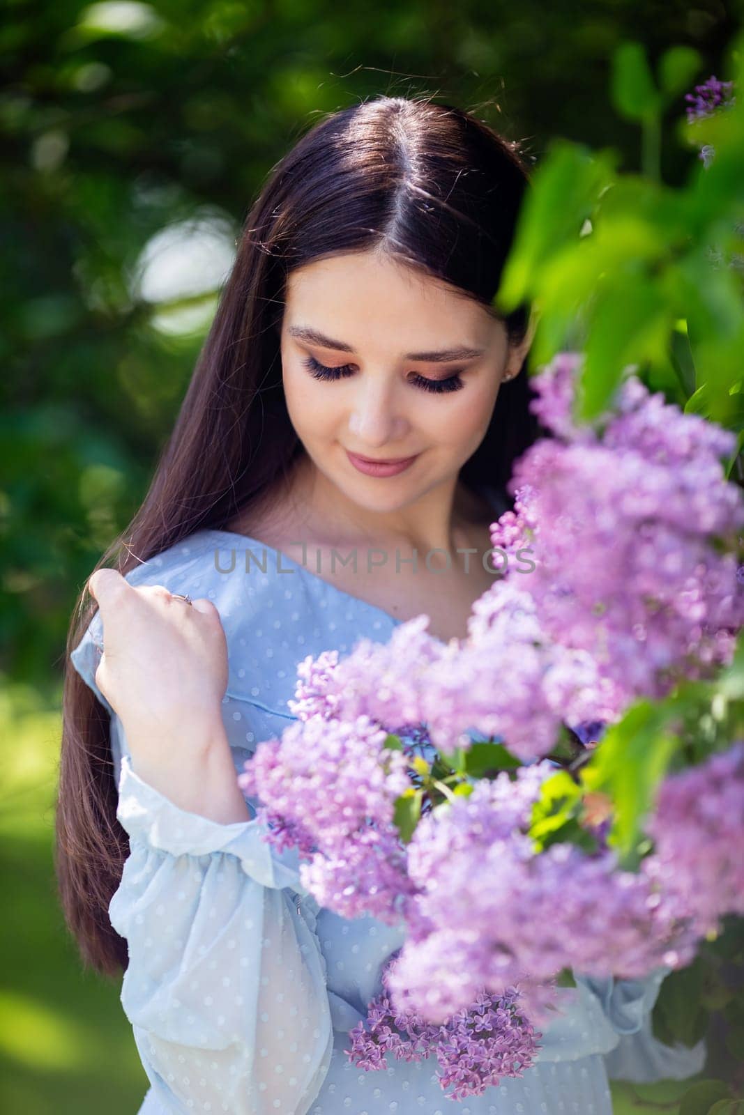 A smiling brunette with long hair stands with lilac flowers, in the spring in the park, on a sunny day. Vertical. Close up