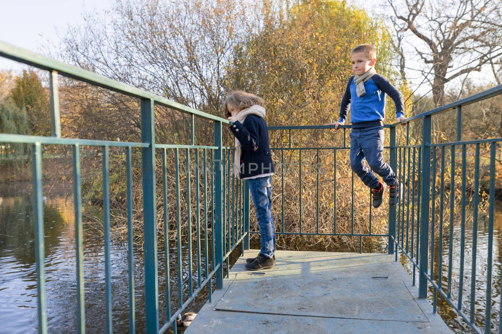 Children boy and girl standing on bridge, looking at ducks, sunny autumn day in park, golden hour