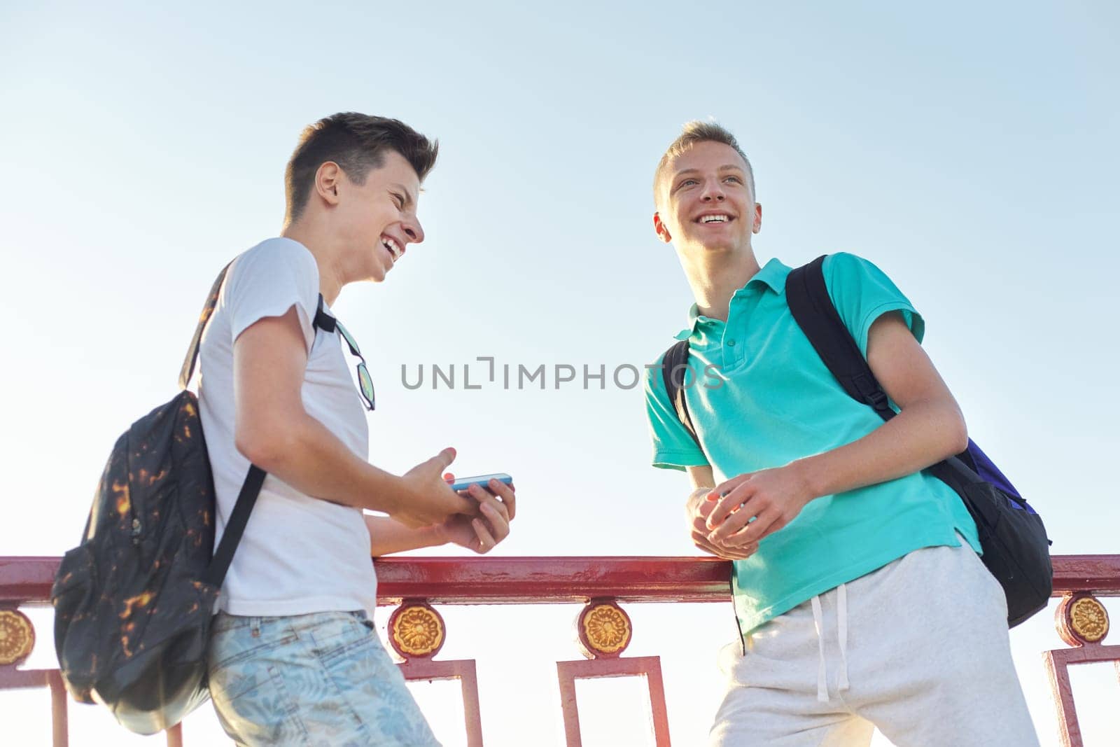 Outdoor portrait of two talking boys teenagers 15, 16 years old, males laughing look at smartphone, golden hour