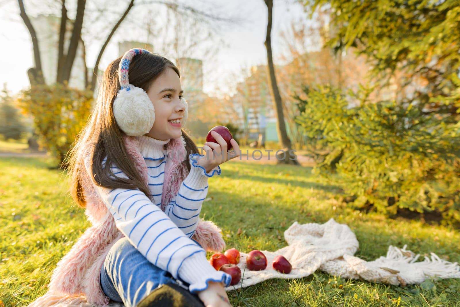 Child girl eating fresh red apples, healthy food in nature, sitting in sunny autumn park, leaf fall golden hour