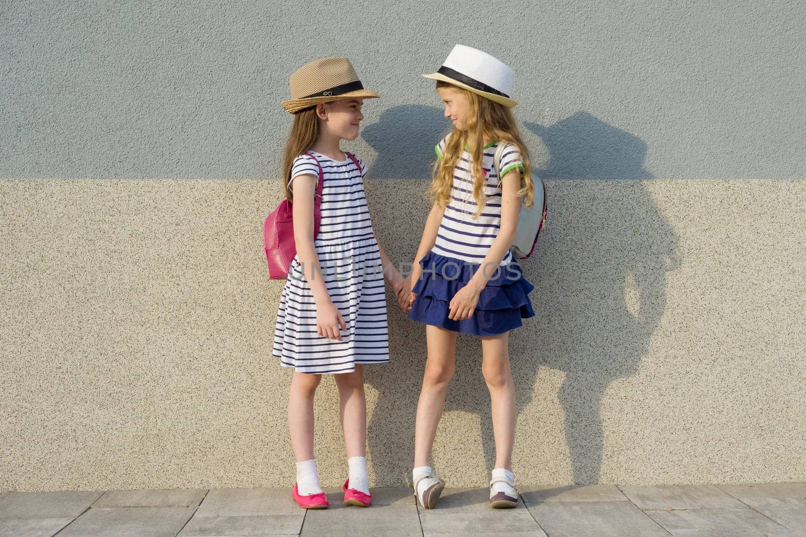 Outdoor summer portrait of two happy girl friends 7,8 years in profile talking and laughing. Girls in striped dresses, hats with backpack, background gray wall