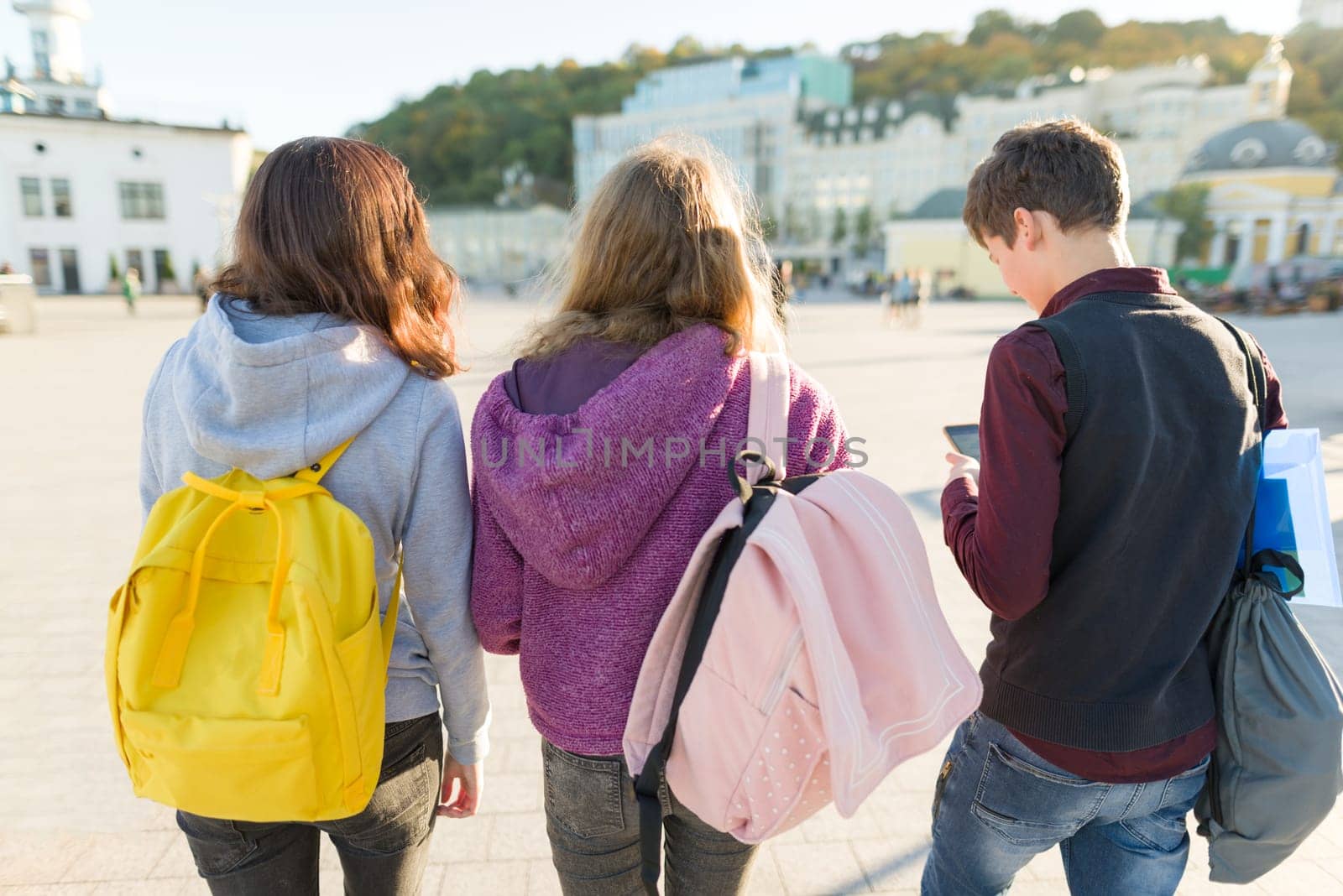 View from the back on three high school students with backpacks. City background, golden hour