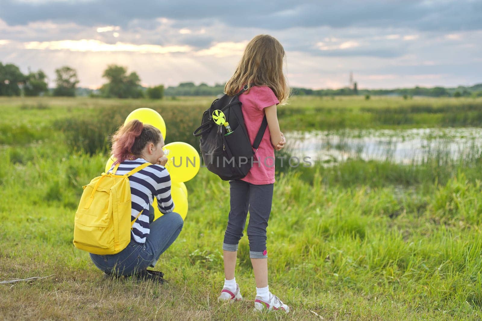Two girls with balloons backpacks back in nature, children near the lake in the meadow, summer evening