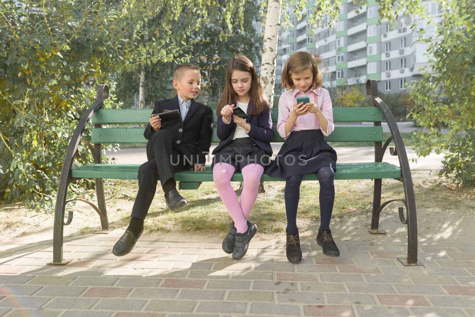 Group of children 7, 8 years old with mobile phones, schoolchildren with backpacks looking into smartphones, outdoor background. Education, friendship, technology and people concept