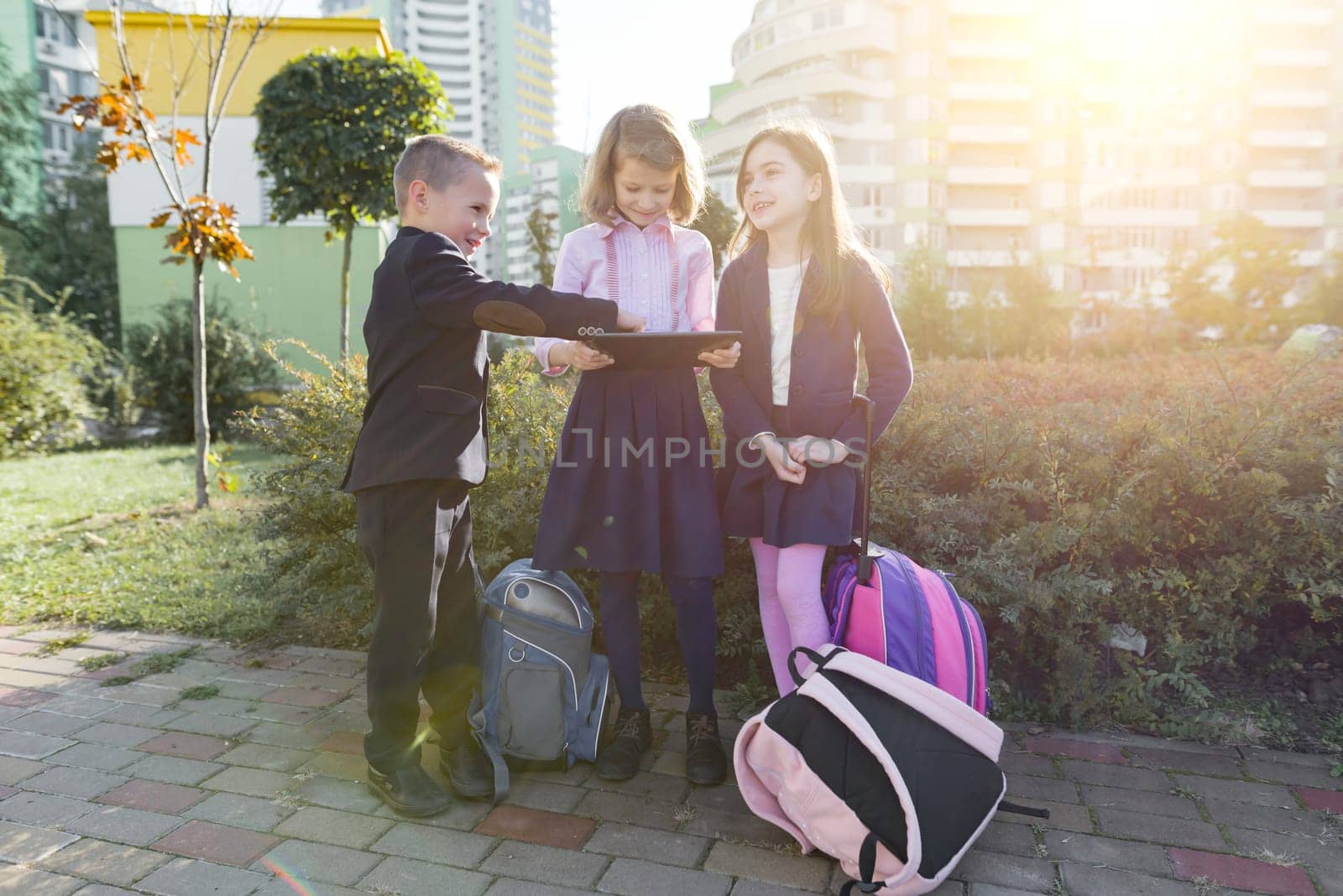 Group of elementary school kids with digital tablet. Outdoor background, children with school backpacks, looking at the tablet, talking. Education, friendship, technology and people concept