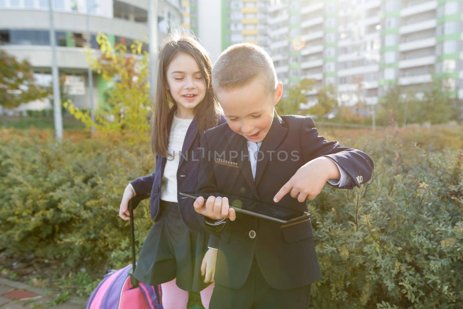 Boy and girl pupils in primary school with digital tablet. Outdoor background, children with school bags, look at the tablet. Education, friendship, technology and people concept