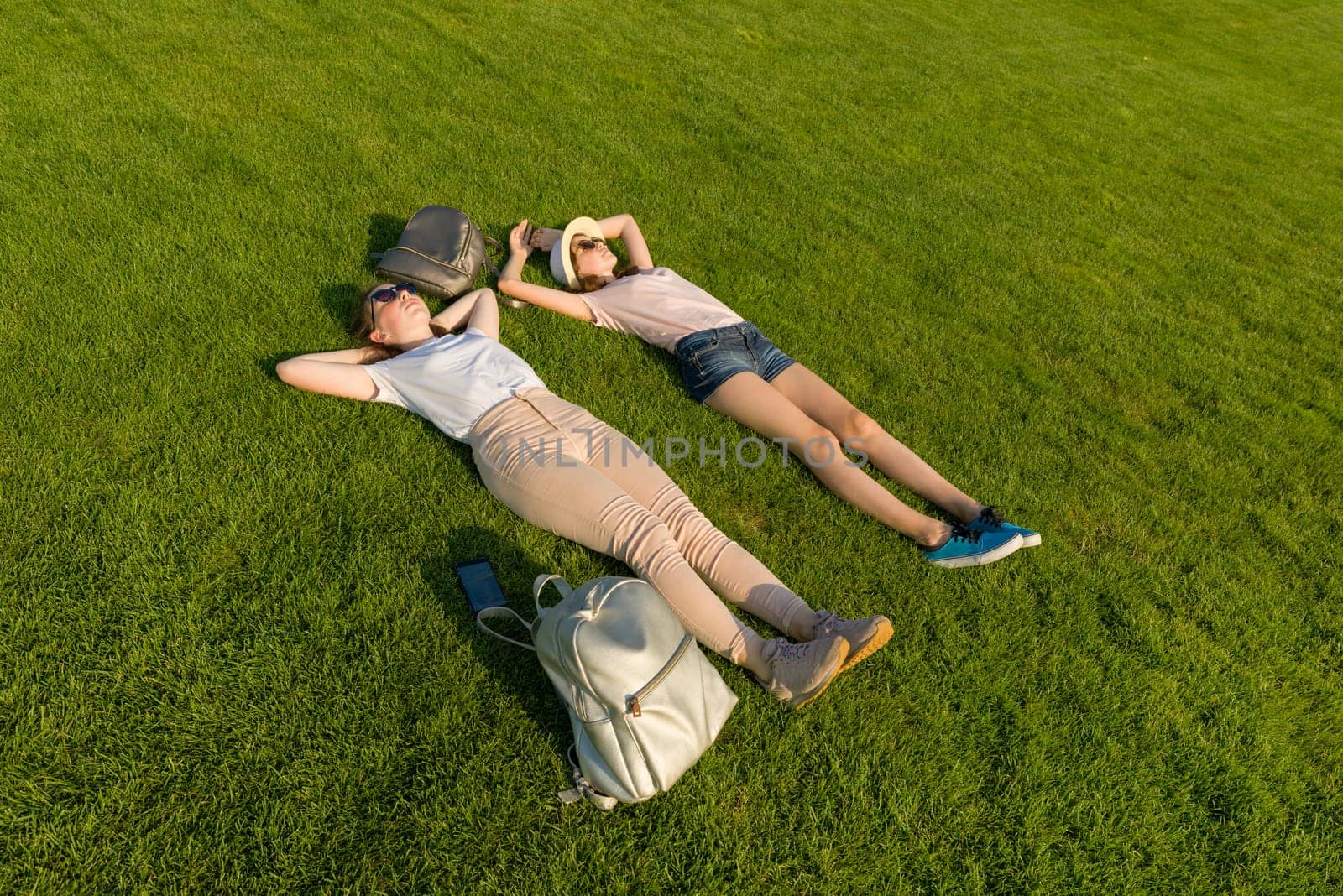 Two young female students with backpacks lying on green lawn grass. by VH-studio