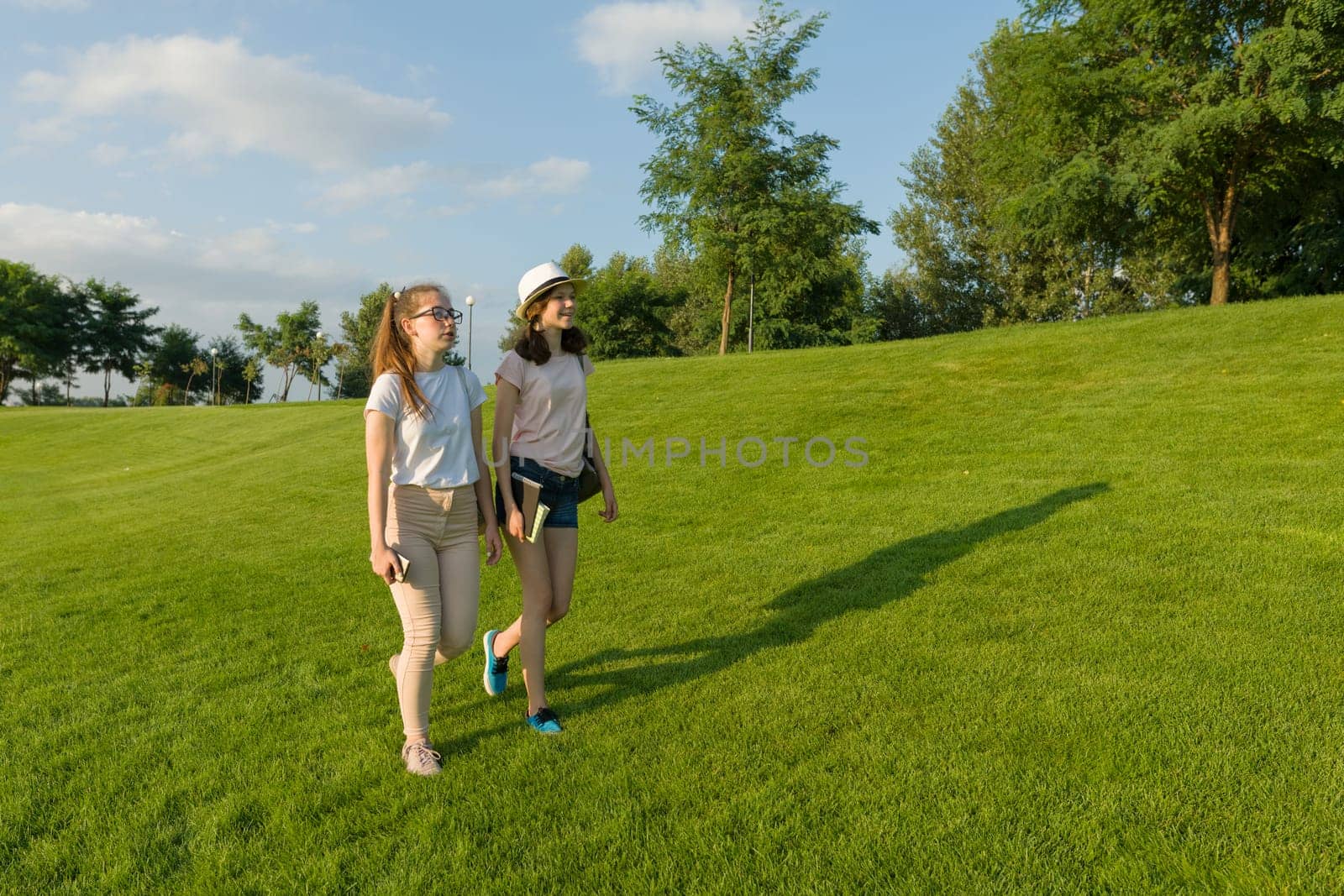 Two teen girls students with backpacks and books walking on green grass in the park, sunny day