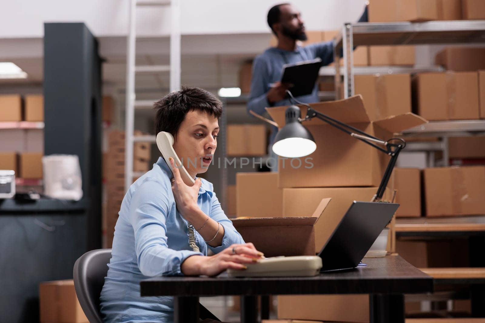 Caucasian manager talking at landline phone with remote supervisor discussing transportation logistics problem while looking at cargo stock on laptop. Employee working at order delivery in warehouse