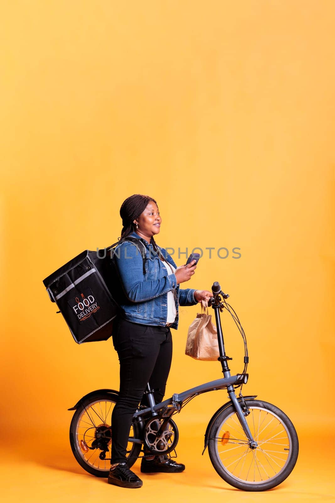 Deliverywoman holding smartphone checking client adreess on fast food app before start delivering lunch orders. Restaurant employee carrying takeout backpack while riding bike as transportation