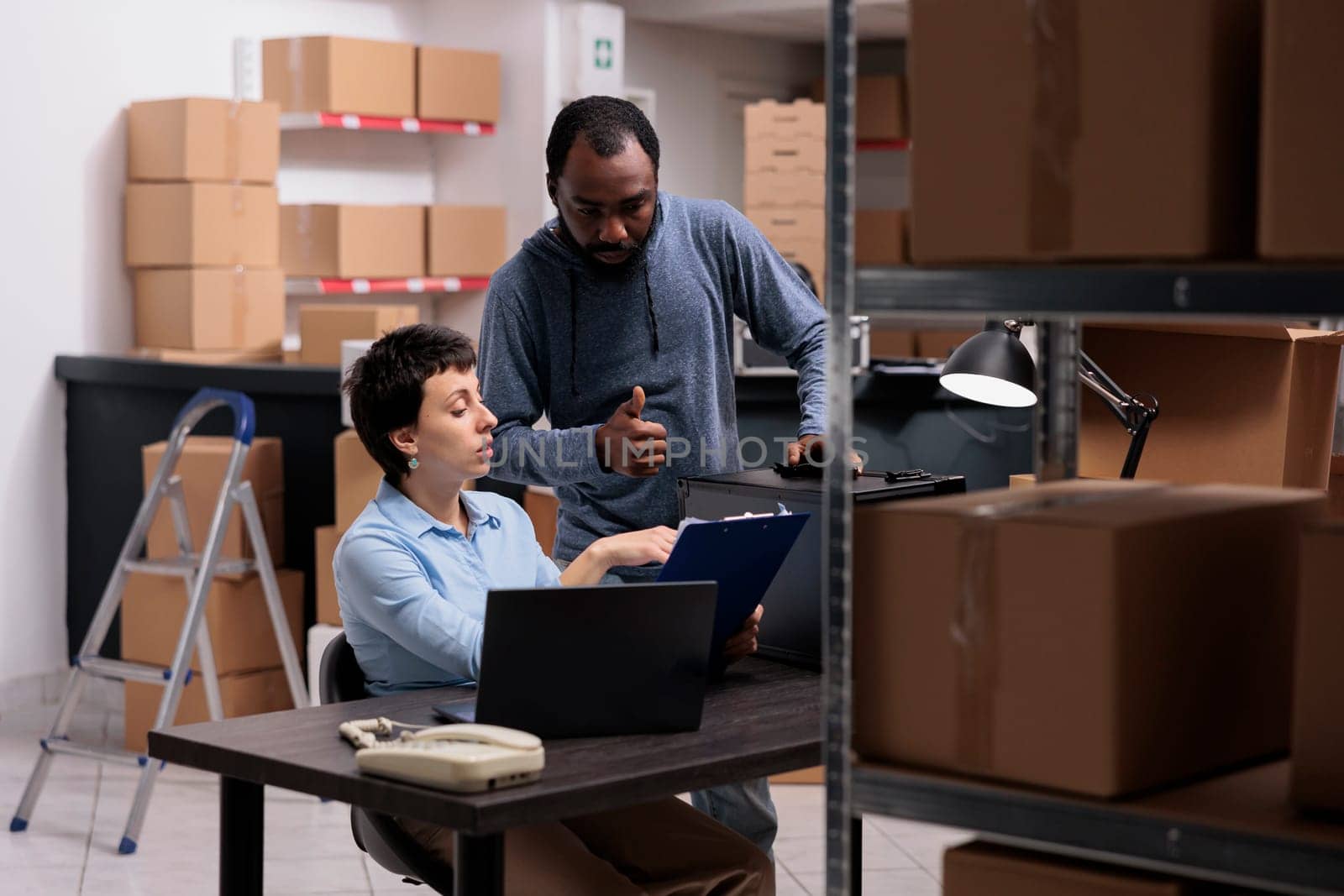 Supervisor woman analyzing metallic delivery order on clipboard discussing transportation logistics with worker while working together in warehouse. Storehouse team checking distribution stock