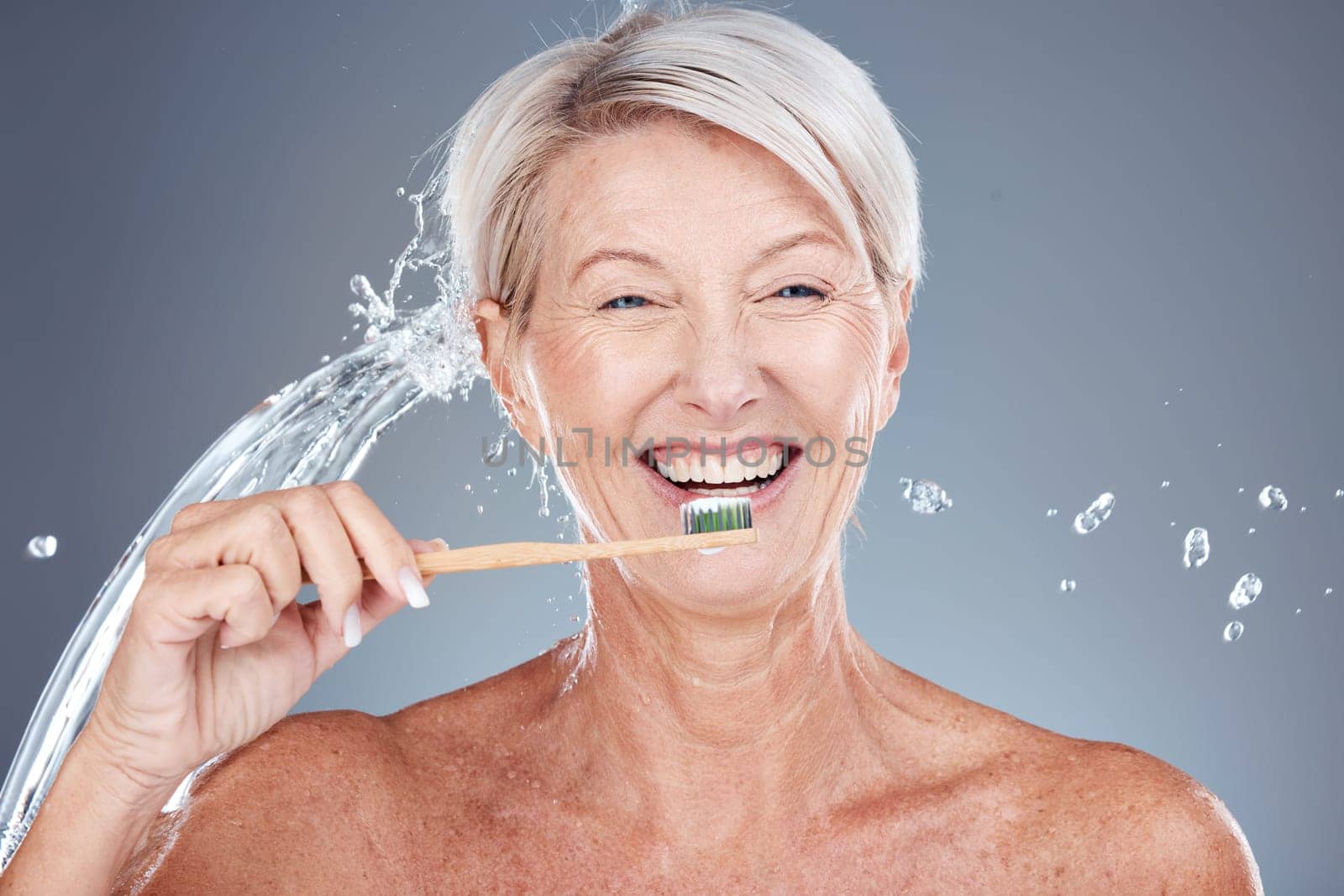 Wellness, water splash and portrait of old woman brushing teeth isolated on gray background in studio. Bamboo, dental care and senior female cleaning teeth with toothbrush for eco friendly hygiene by YuriArcurs