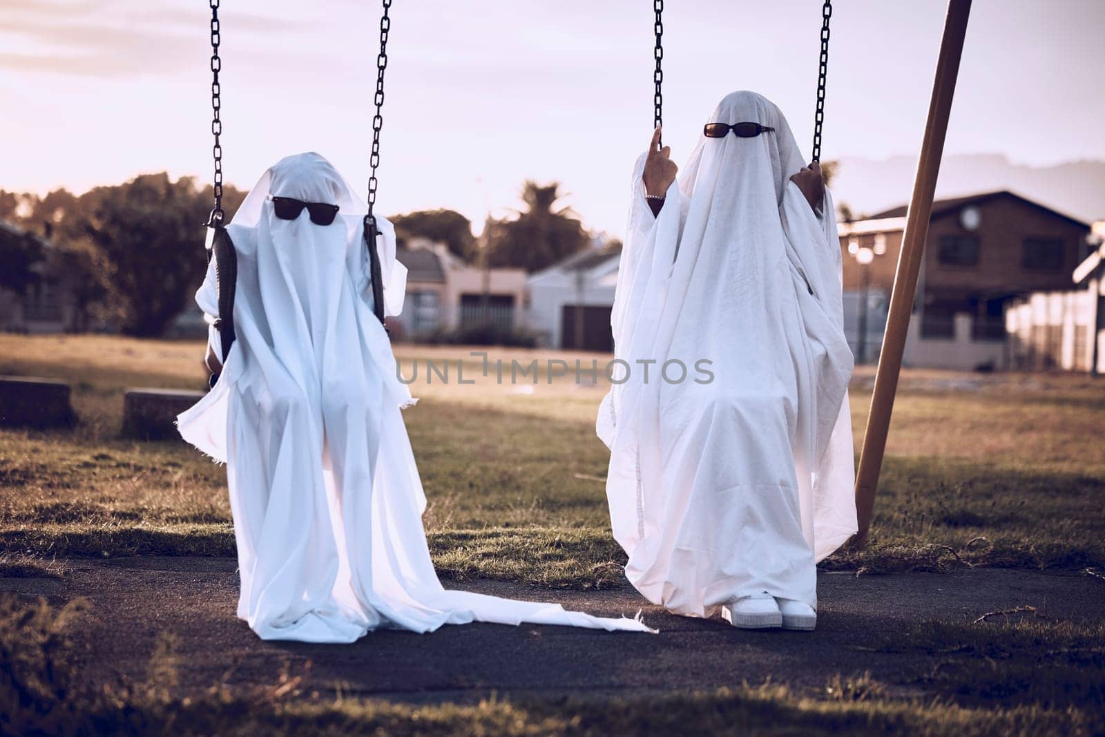 Park, halloween and friends in a ghost costume in a kids playground in celebration of a scary fantasy holiday. Glasses, creepy and crazy couple in horror, creative and spooky white sheets on a swing.