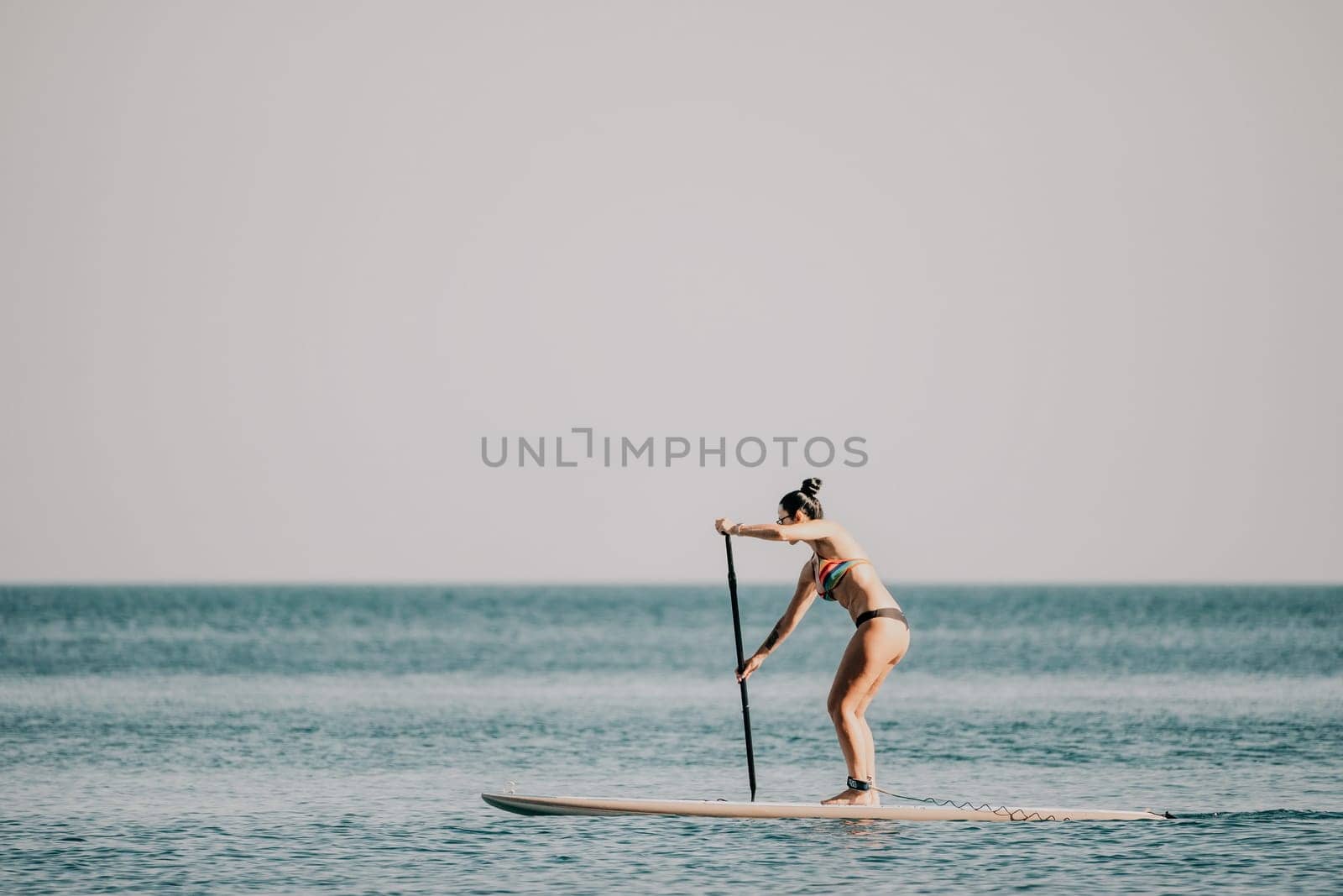 Sea woman sup. Silhouette of happy middle aged woman in rainbow bikini, surfing on SUP board, confident paddling through water surface. Idyllic sunset. Active lifestyle at sea or river