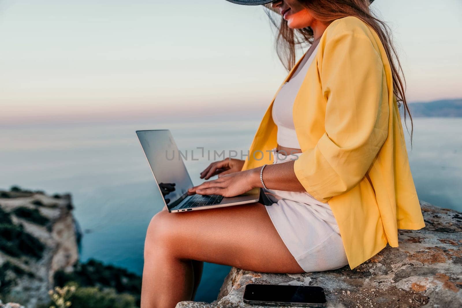 Successful business woman in yellow hat working on laptop by the sea. Pretty lady typing on computer at summer day outdoors. Freelance, travel and holidays concept.