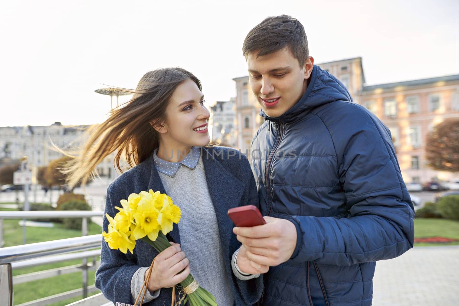 Outdoor portrait of beautiful romantic couple, young man and woman with bouquet of yellow flowers of daffodils and looking in smartphone, spring city background.