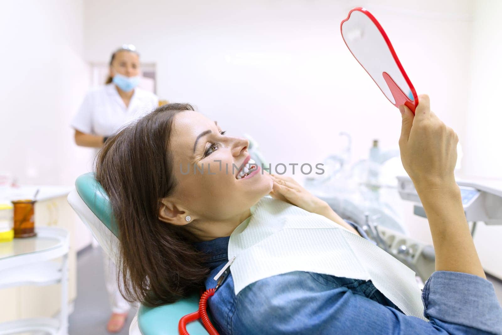 Female patient looking at her teeth in mirror while sitting in dental chair. by VH-studio