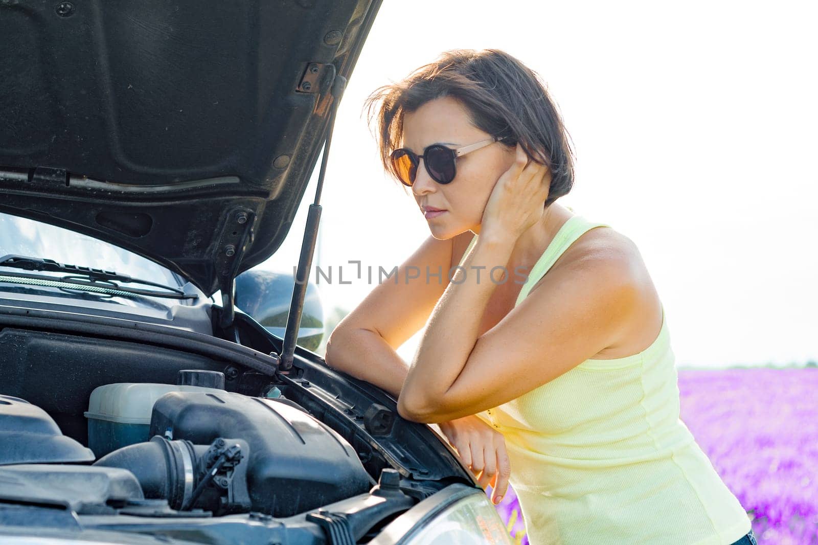 Distressed woman driver standing near broken car, raised hood. Summer nature background, lavender field.