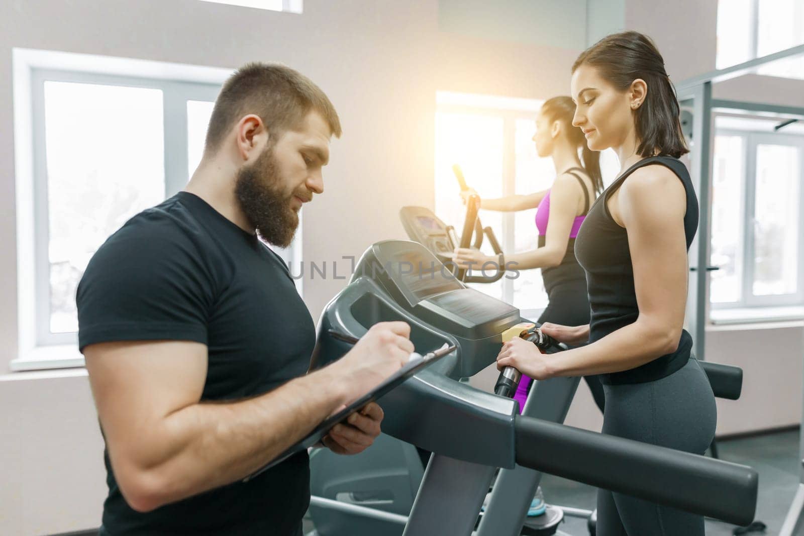 Young smiling fitness women with personal trainer an adult athletic man on treadmill in the gym. Sport, teamwork, training, healthy lifestyle concept.