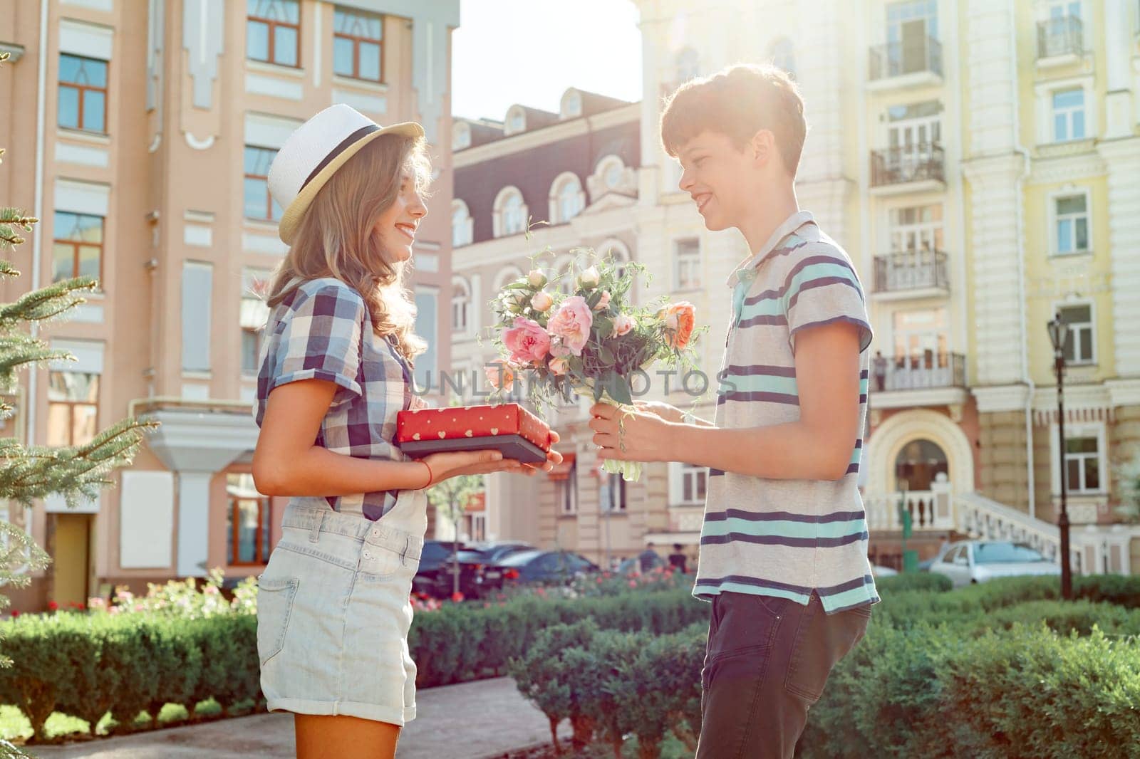 Teen boy congratulates girl with bouquet of flowers and gift, outdoor portrait couple happy youth.