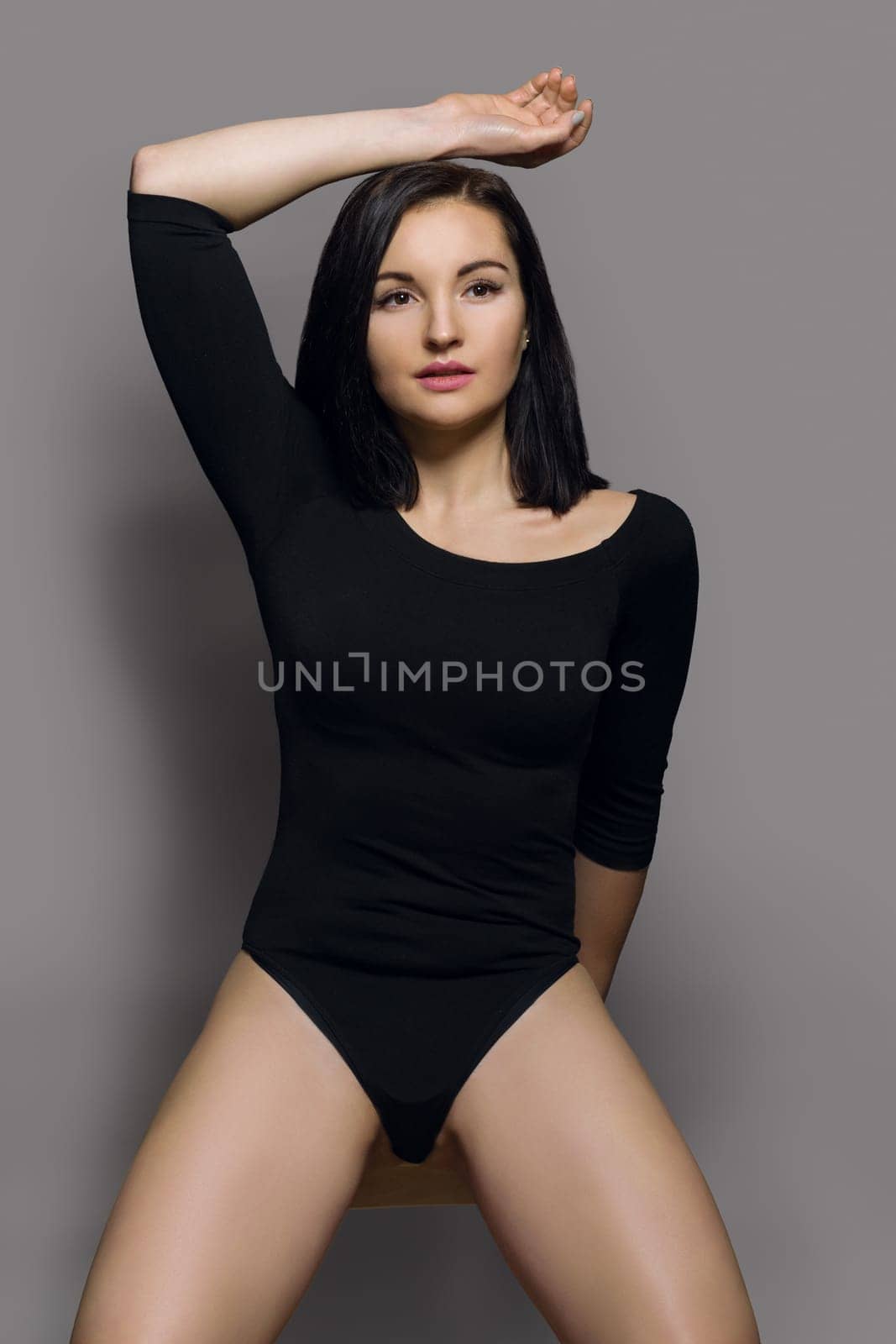 Beauty fashion portrait of young beautiful woman in black bodysuit with long sleeves. Gray studio background, perfect makeup, healthy hair, athletic body