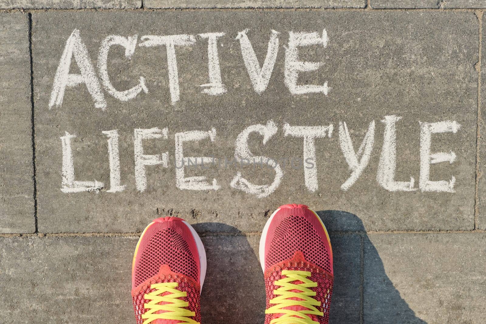 Text active lifestyle written on gray pavement with woman legs in sneakers, view from above.