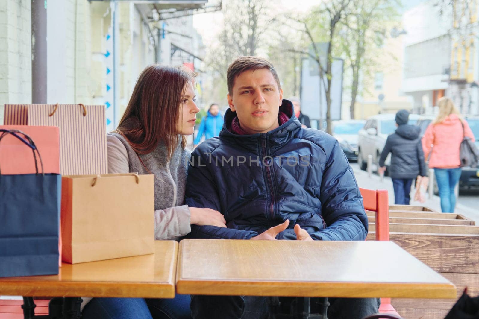 Young tired couple with shopping bags in spring street cafe, waiting for cup of coffee and tea