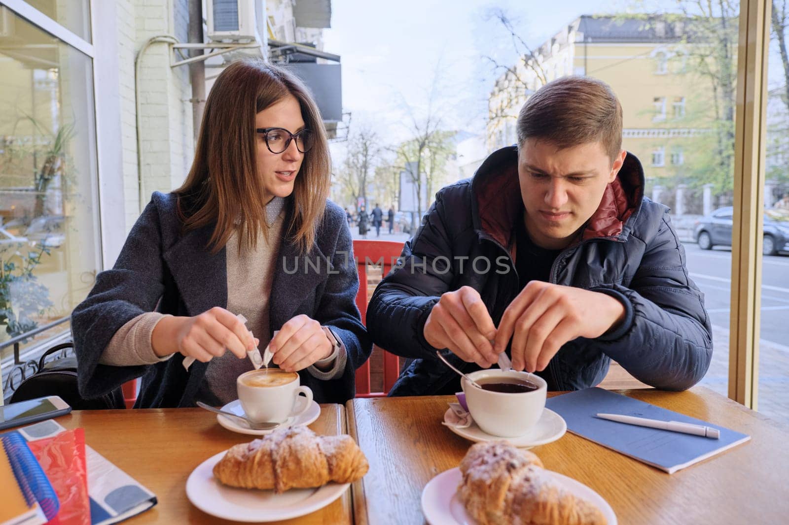 Young male and female friends students sitting in outdoor cafe, talking, drinking coffee, tea, eating croissants. On table textbooks, notebooks, city background