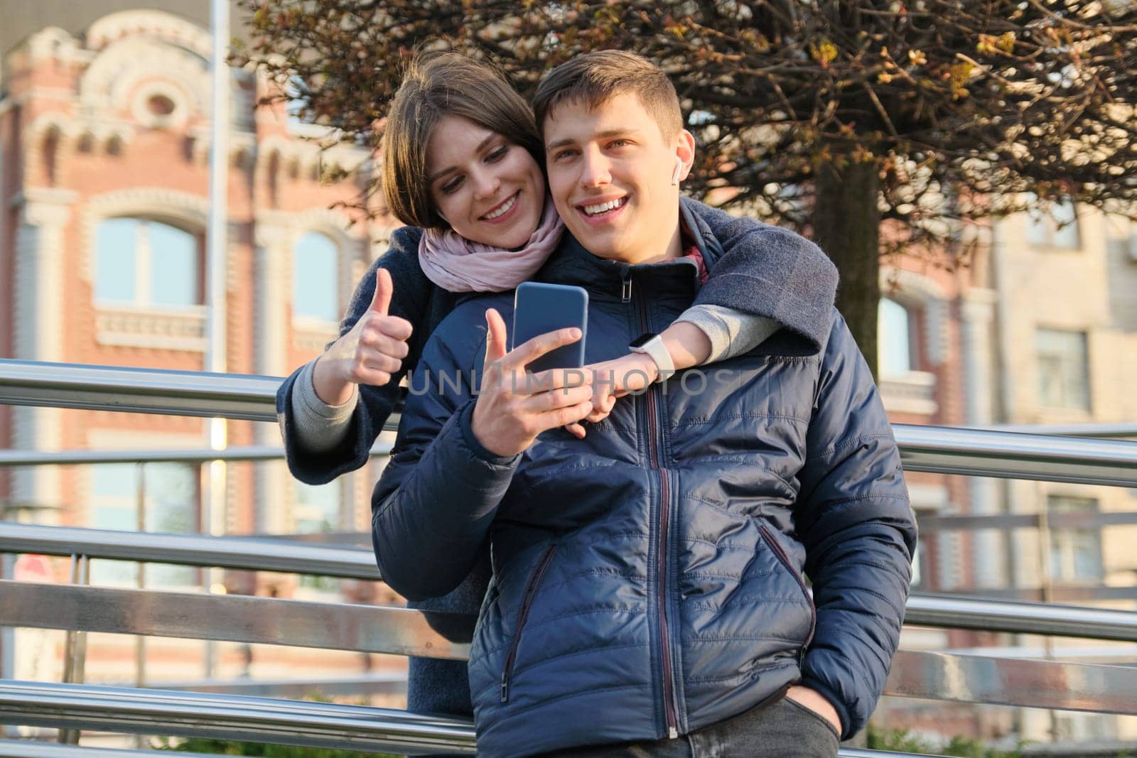 Outdoor portrait of couple showing gesture sign ok. Young man and woman with smartphone in headphones, city background golden hour.