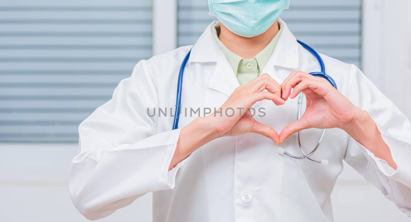 Closeup of doctor woman wearing white coat standing making heart finger shape by Sorapop